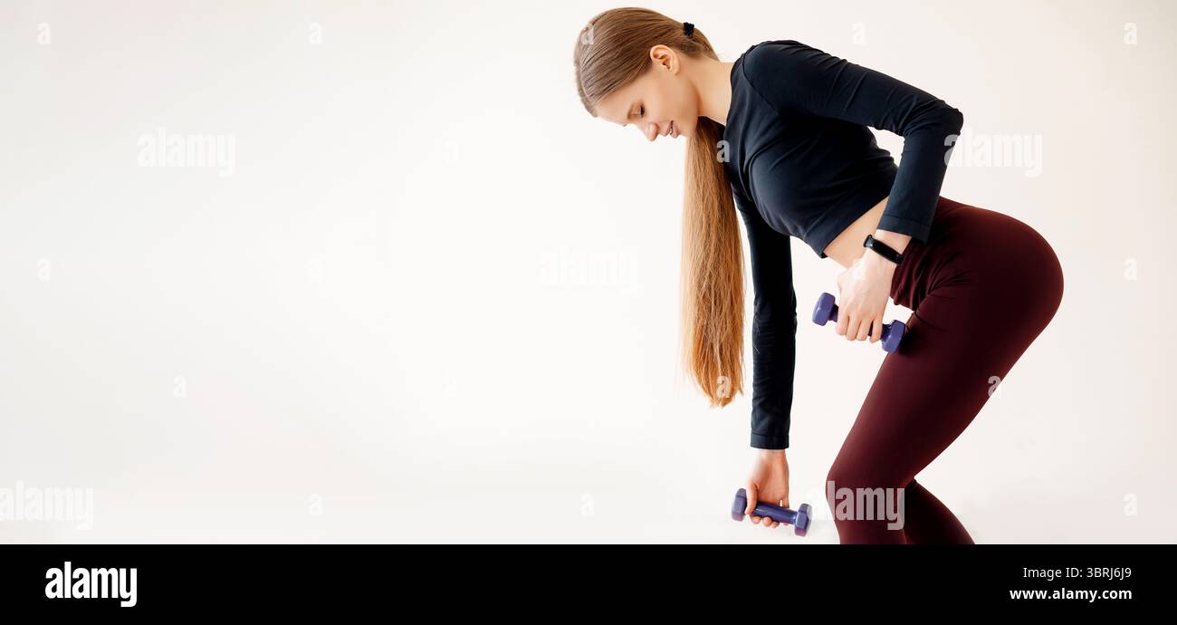 Schlanke Frau mit perfekter Körperform, die regelmäßiges Workout mit Sportgeräten in Innenräumen macht. Fitte Frau trainiert mit Kurzhanteln und übt rumänisch d Stockfoto
