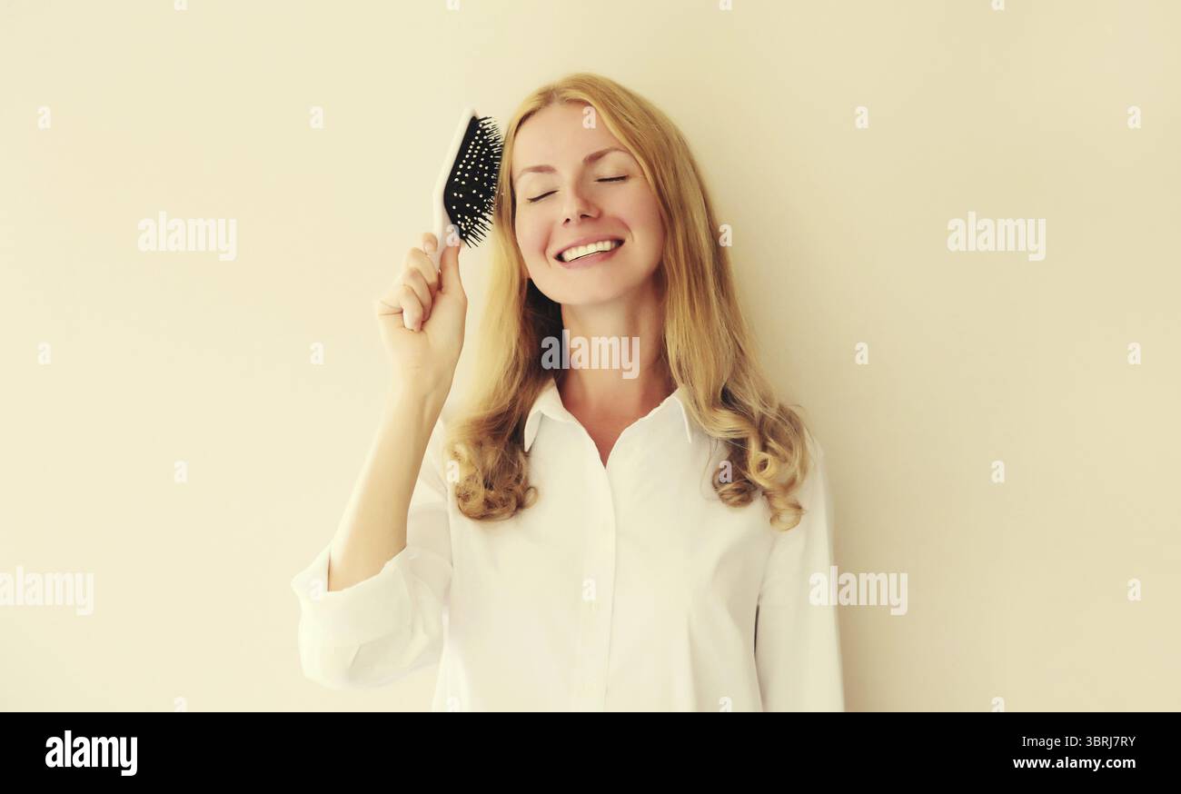 Porträt einer glücklichen lächelnden kaukasischen blonden jungen Frau, die ihr Haar mit Kamm auf weißem Studiohintergrund kämmt Stockfoto
