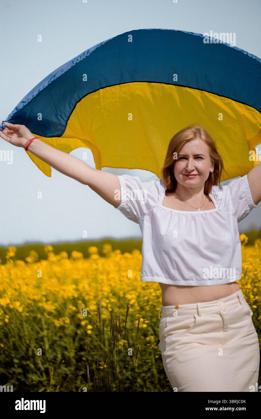 Die fröhliche Erwachsene Frau winkt die ukrainische Flagge über ihrem Kopf während der Nationalfeier auf einem Rapsfeld Stockfoto