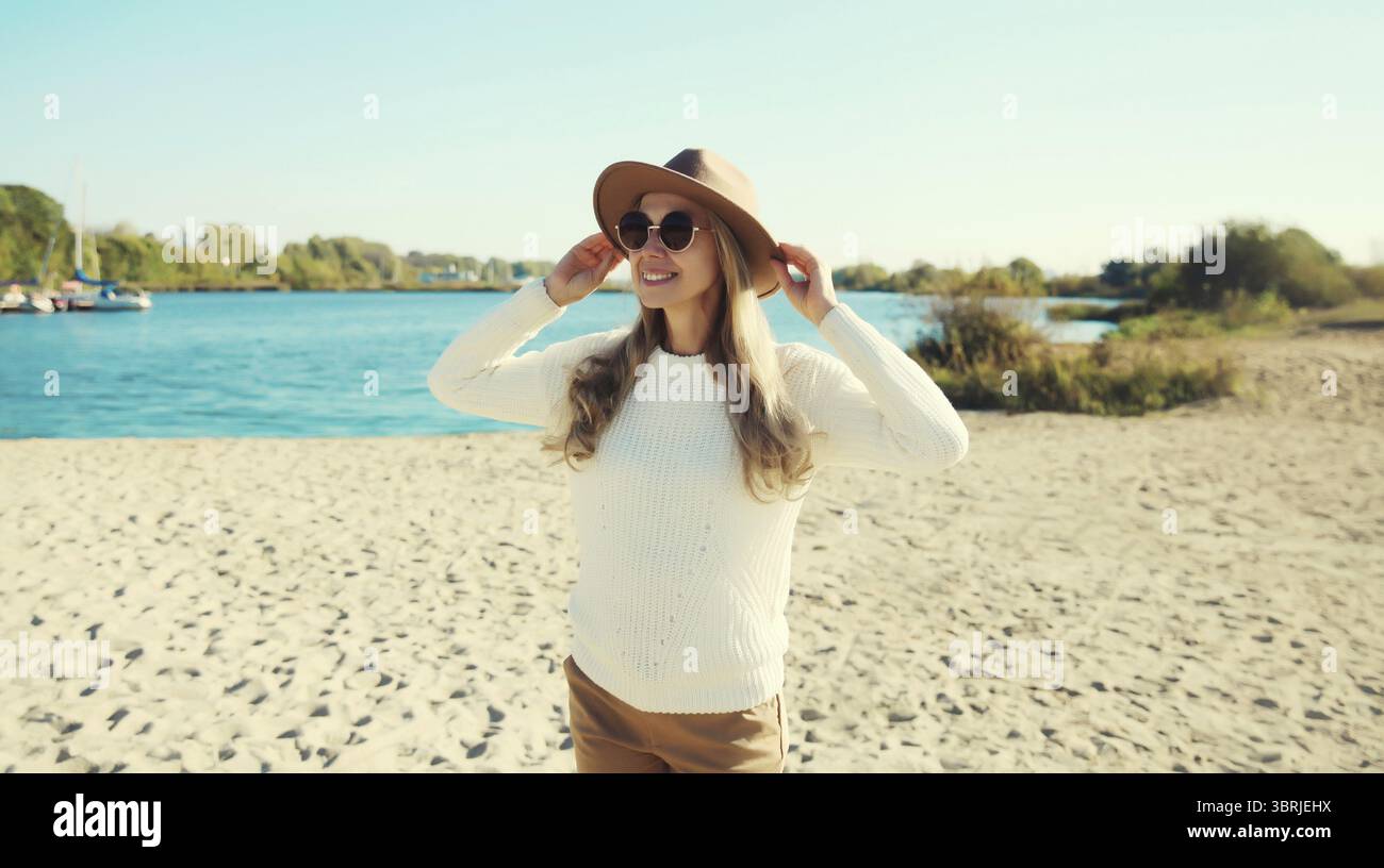 Stilvolle glückliche Frau, die am sonnigen Strand steht, warmes Wetter, lächelt, wegschaut, Hut tragen, Brille, weißer Pullover, auf Meeresküste Hintergrund Stockfoto