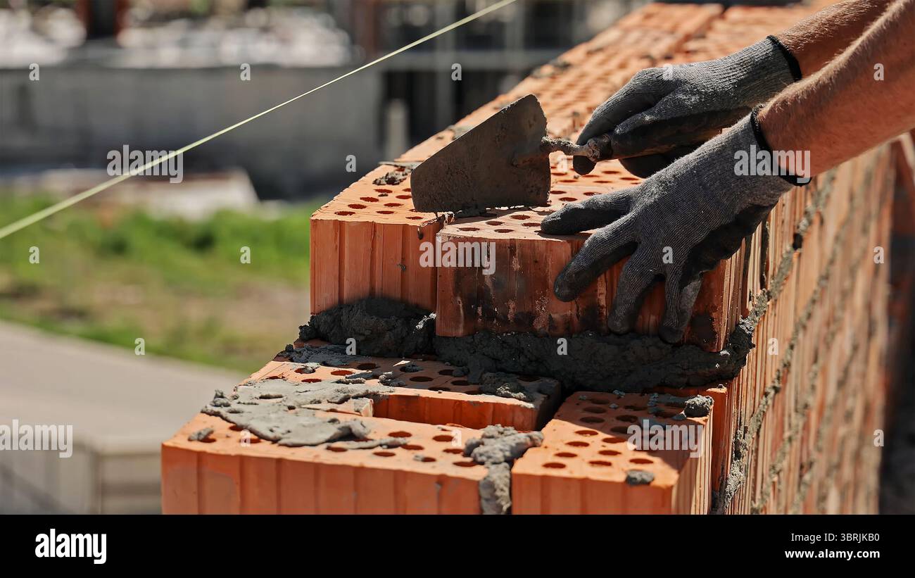 Maurer, der Ziegelsteine mit Zementmörtel legt. Nahaufnahme eines Bauarbeiters mit Handschuhen, der mit einer Kelle rote Lochziegel mit nassem Zement legt Stockfoto