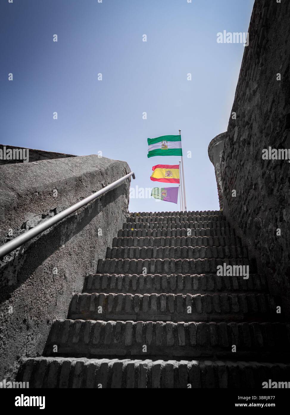 Mit Blick auf die Steintreppe in Richtung Himmel winken drei Fahnen stolz über Málaga: Andalusien, Spanien und das Wahrzeichen der Stadt. Stockfoto
