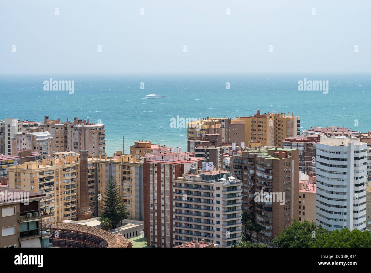 Ein ruhiger Blick auf die Küste von Málaga mit einer einzigen weißen Yacht, die auf türkisfarbenem Wasser jenseits der Skyline des Hochhauses driftet. Stockfoto