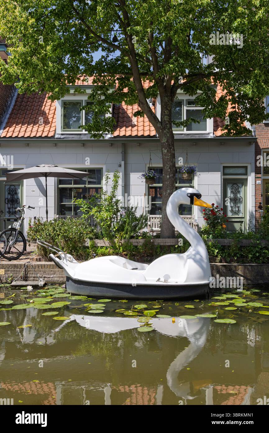 Ein entzückendes Schwan-Paddelboot ruht auf einem ruhigen niederländischen Kanal in Delft und spiegelt bezaubernde Häuser am Wasser und üppiges Grün unter einem klaren Himmel wider. Stockfoto