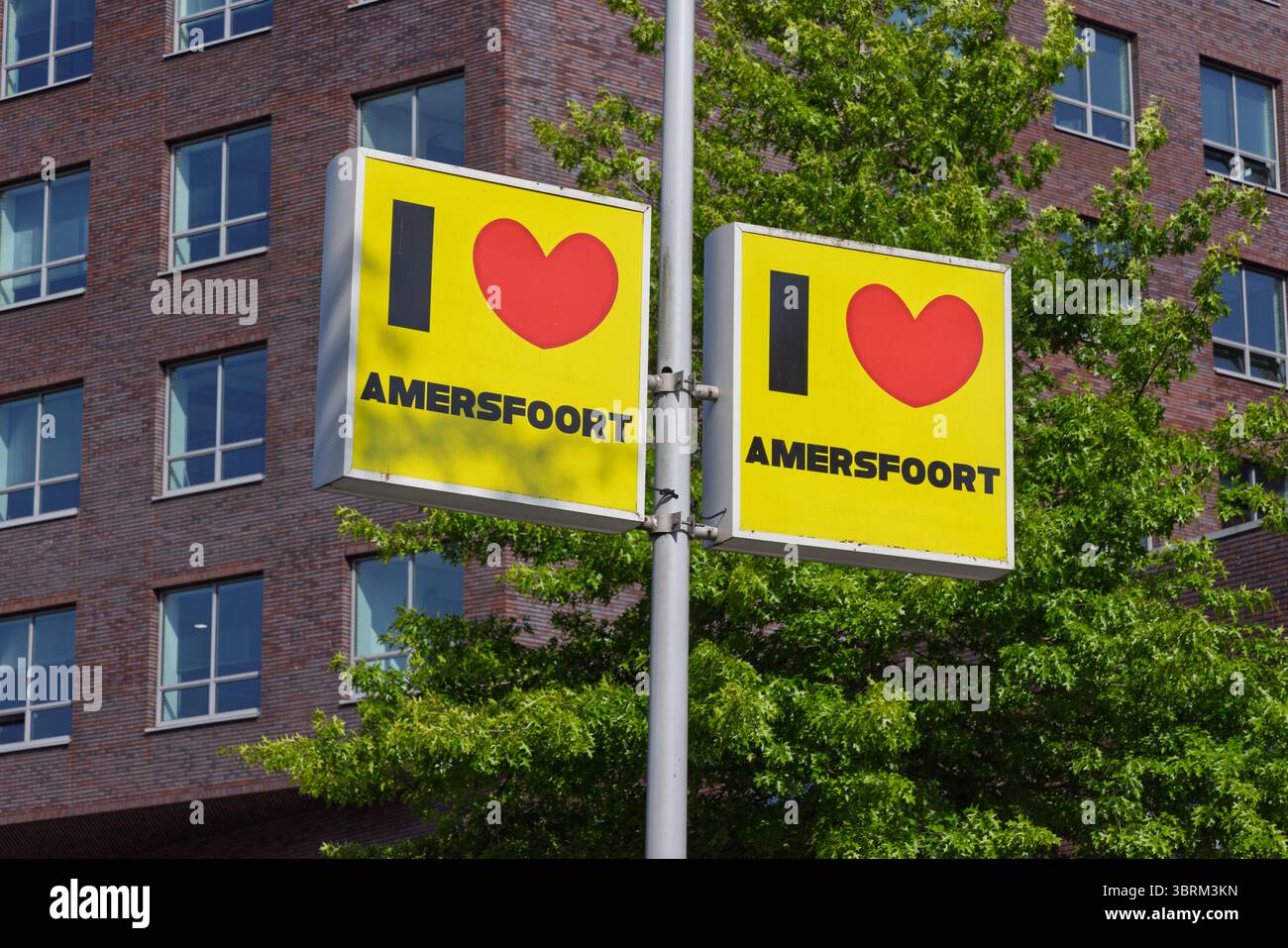 Städtisches Straßenschild im auffälligen gelben „I love Amersfoort“-Design, das den Stolz der Stadt in Amersfoort, Niederlande, mit modernem Gebäude und Bäumen auf der Rückseite zum Ausdruck bringt Stockfoto