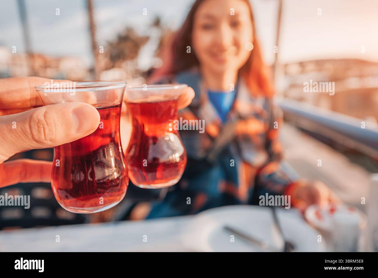 Zwei türkische Teegläser stoßen bei einem Toast an, während ein lächelnder Tourist den Moment vor einer lebhaften Kulisse bei Sonnenuntergang genießt Stockfoto