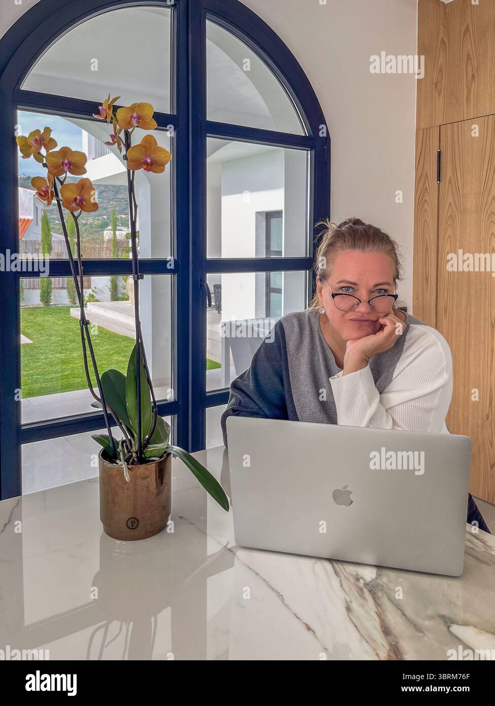 Faro, Portugal - 13. Juli 2025: Frau, die in einem hellen Home Office mit großen Fenstern und einer eleganten Orchideenpflanze an einem Apple-Laptop arbeitet Stockfoto