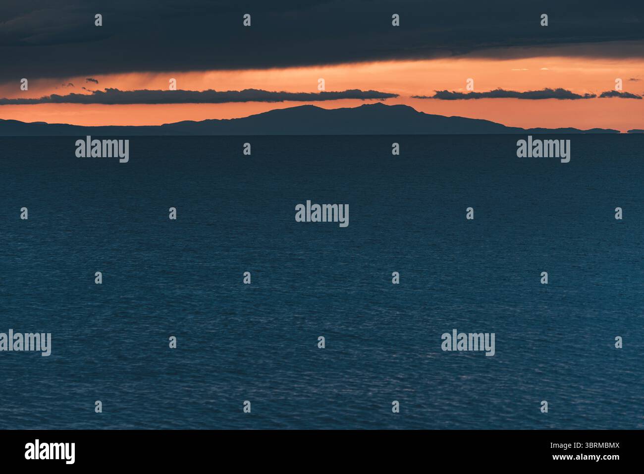 Dramatische Orangetöne, die den Himmel während des Sonnenuntergangs bemalen und Reflexionen über einem dunkelblauen Meer mit weit entfernten Inseln und Wolken werfen Stockfoto