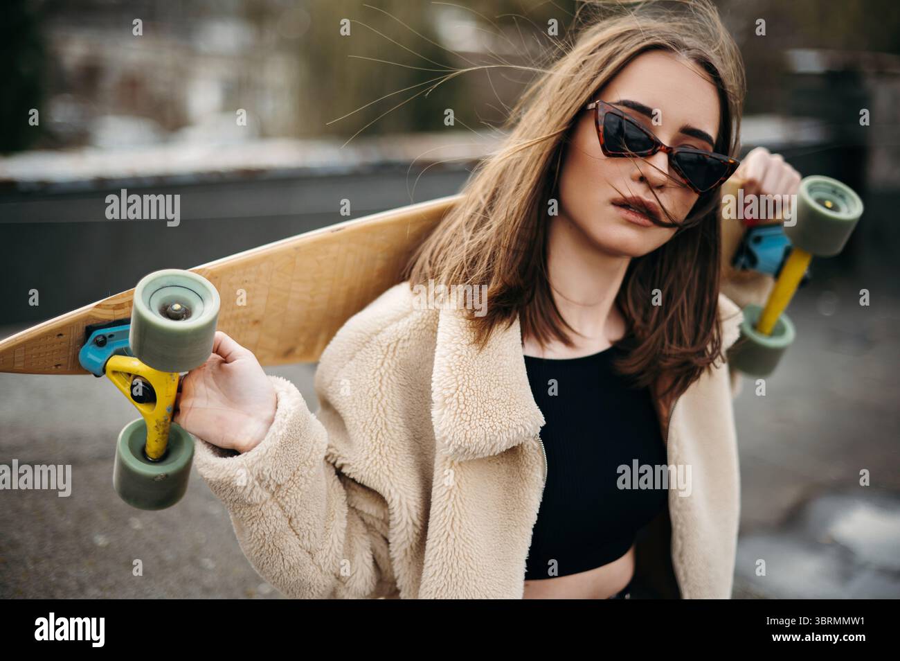 Junge Brünette mit professionellem Skateboard auf den Schultern, die Zeit auf dem Dach des hohen Gebäudes verbringt. Stilvolle Frau trägt legere Kleidung und moderne Bungl Stockfoto
