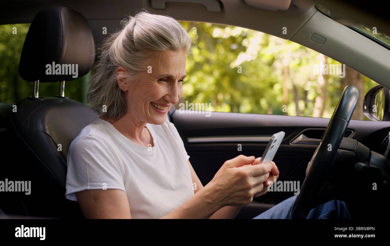 Fröhliche kaukasische reife Frau ältere ältere ältere Frau Rentner Auto Fahrer im Auto mit Handy spielen Spiel Ballen Faust Stockfoto