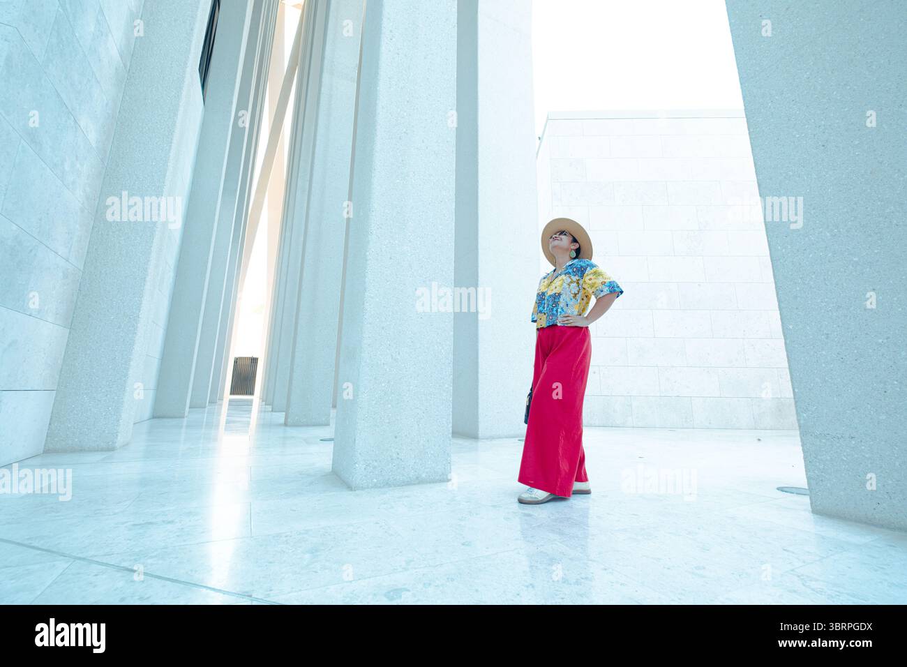 Eine Frau steht zwischen hohen weißen Strukturen, trägt eine bunte Bluse, eine rote Hose und einen Hut mit breiter Krempe und blickt nach oben Stockfoto