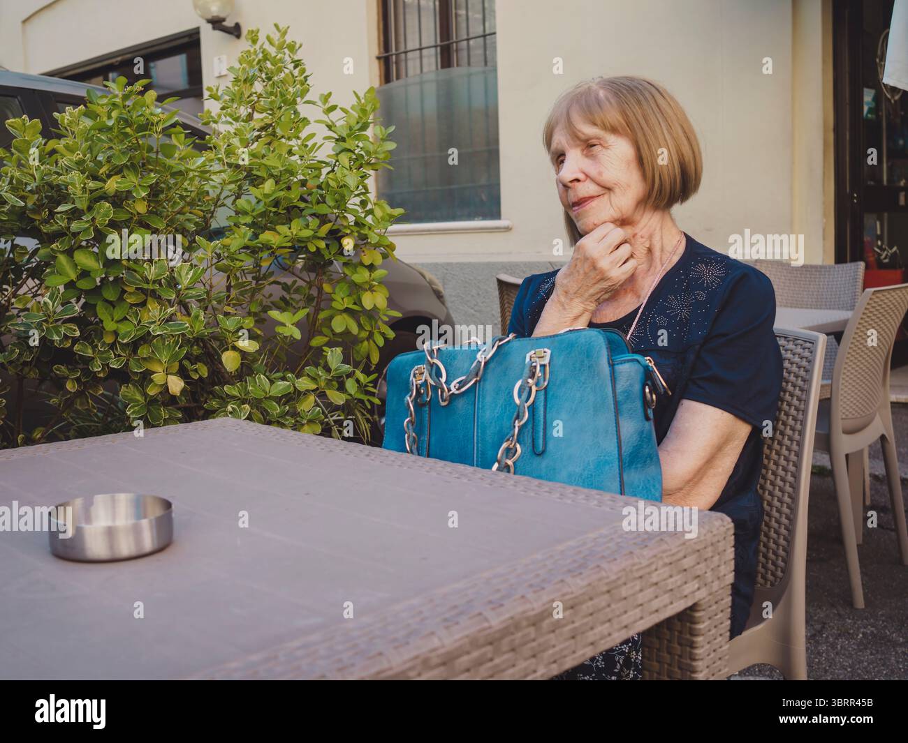 Nachdenkliche Seniorin mit kurzen blonden Haaren und einer blauen Decke, die an einem Kaffeetisch im Freien sitzt, Hand am Kinn und mit einem grünen Busch im Hintergras wegblickt Stockfoto