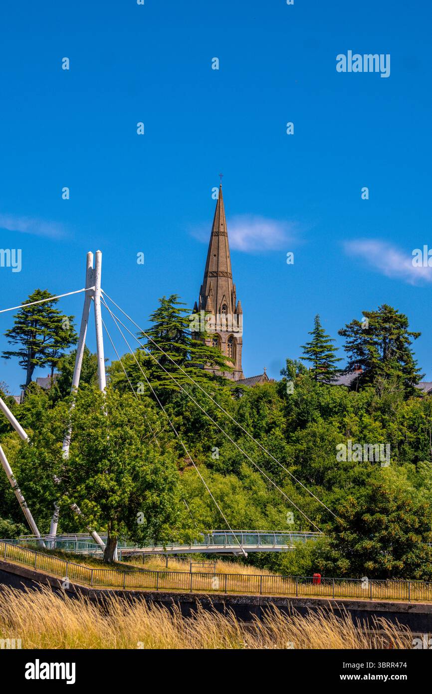 Millers überquert die Fußgängerbrücke über den Fluss exe in Exeter, Devon, mit einem Kirchturm, der an einem hellen Sommertag über den Baumkronen ragt Stockfoto