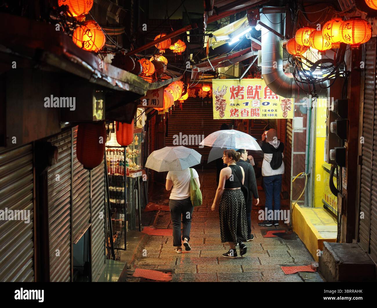 Jiufen, Taiwan - 8. November 2024 - Touristen mit Sonnenschirmen spazieren durch eine enge, nasse Gasse, die von traditionellen roten Laternen und Lampen beleuchtet wird i Stockfoto