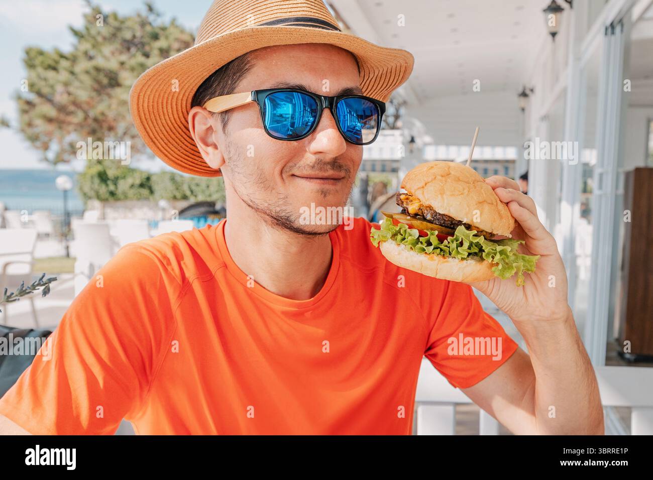 Junger Mann mit Sonnenbrille und Strohhut, der einen Burger in einem Restaurant im Freien mit Meerblick hält Stockfoto