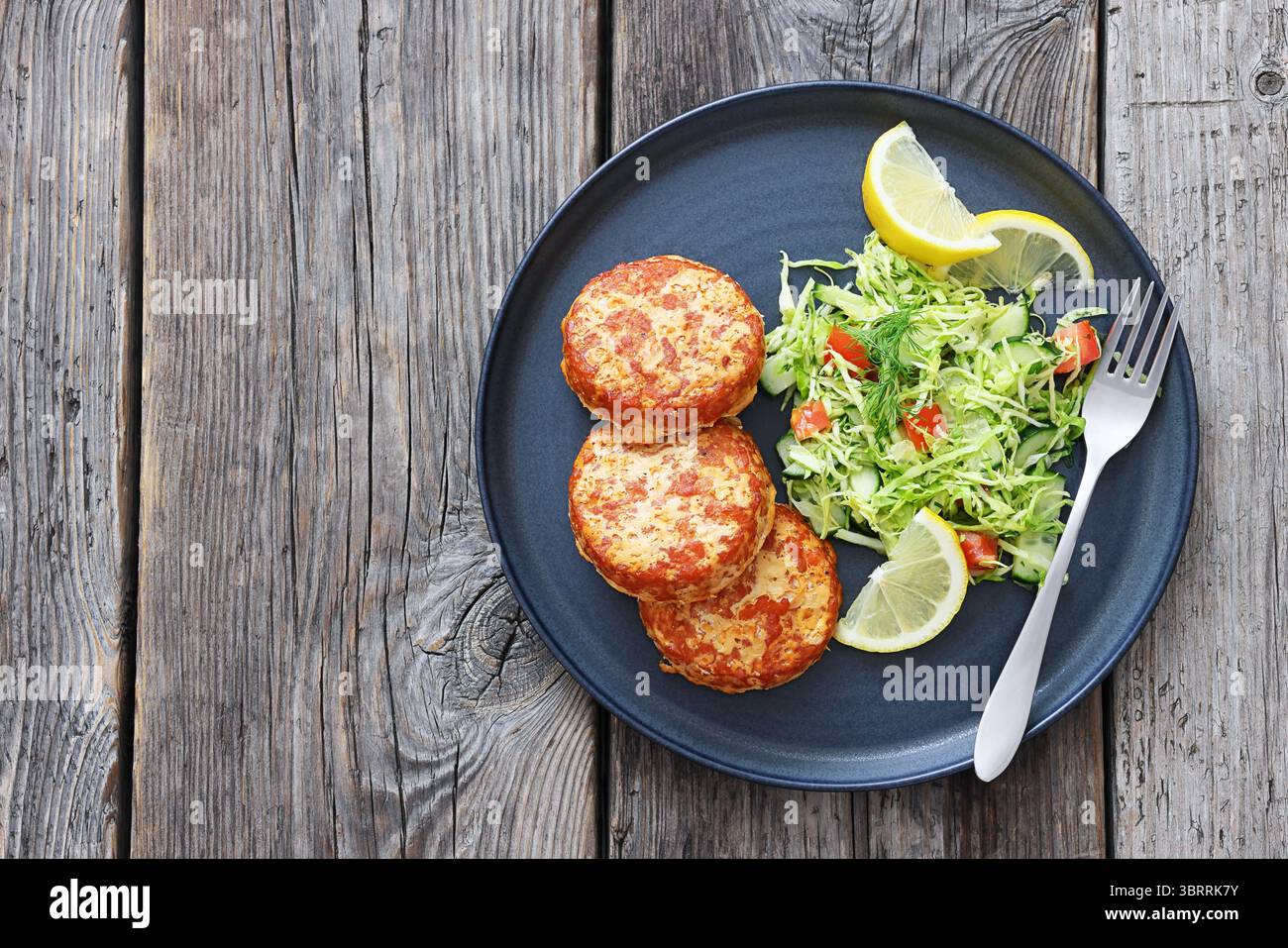 Geräucherte Lachsburger, Pasteten oder Kuchen serviert mit Krautsalat und Zitronenscheiben auf einem Teller mit Gabel auf rustikalem Holztisch, Blick von oben, flacher Lay, kostenloses Spa Stockfoto