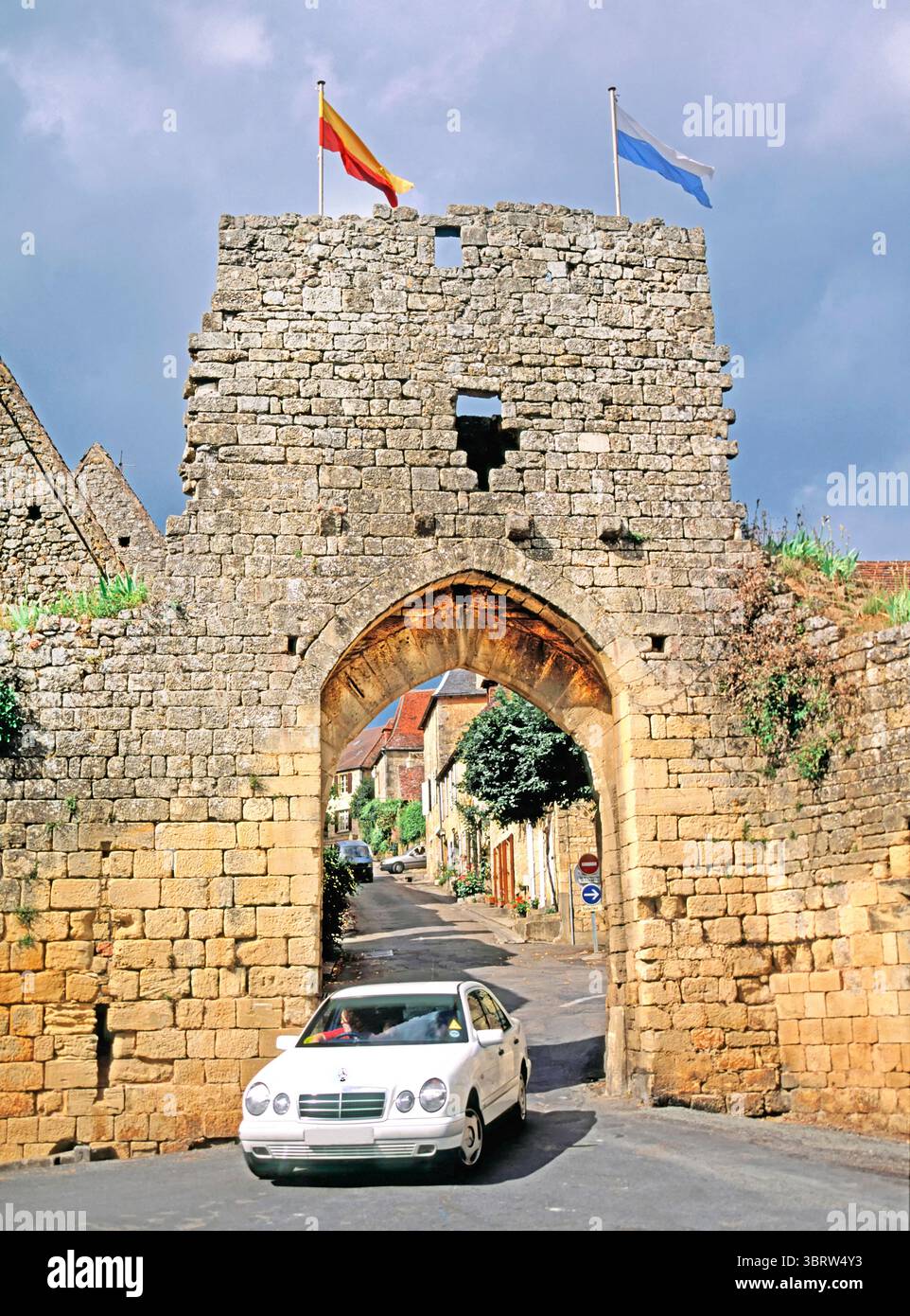 Domme Dordogne Porte Del Bos Archival der 1990er Jahre sonniger Tagesblick eines weißen Mercedes-Autos, der durch einen Bogen in einer Steinmauer und ein Tor fährt, in einer Straßenszene aus den 90er Jahren, eingerahmt von einer Bogenlandschaft der schmalen mittelalterlichen Straße dahinter mit zwei Flaggen vor allem im Département Dordogne in der Region Nouvelle-Aquitaine im Südwesten Frankreichs. Stockfoto