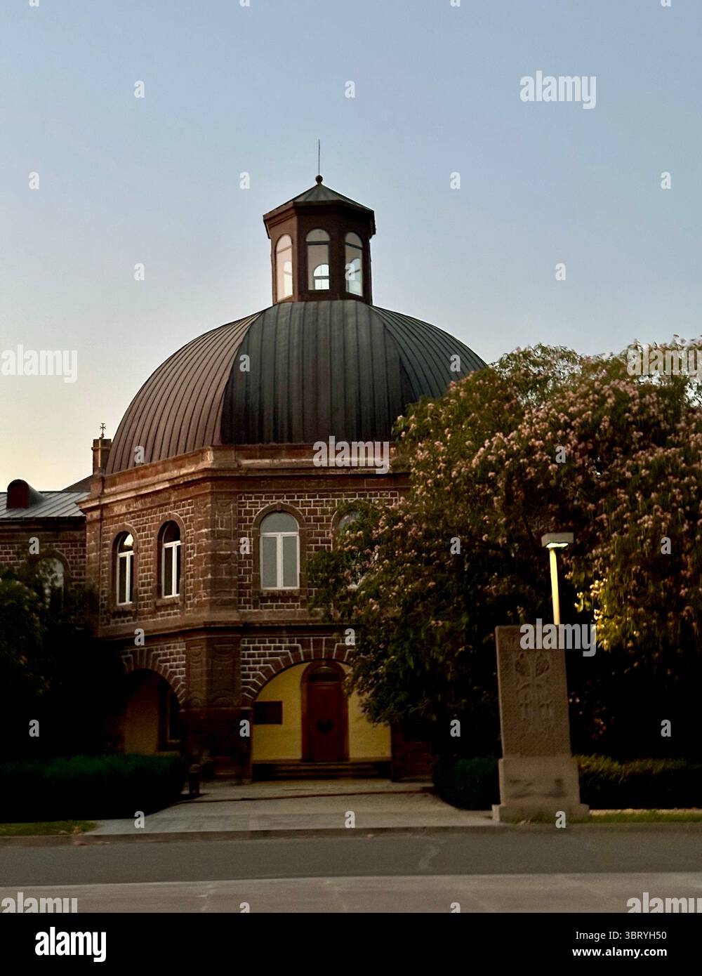 Historisches Steingebäude mit einem Kuppeldach, das bei Sonnenuntergang gefangen gehalten wird, umgeben von Bäumen und einem traditionellen armenischen Khatschkar. - Smartphone-aufgenommenes Stockfoto