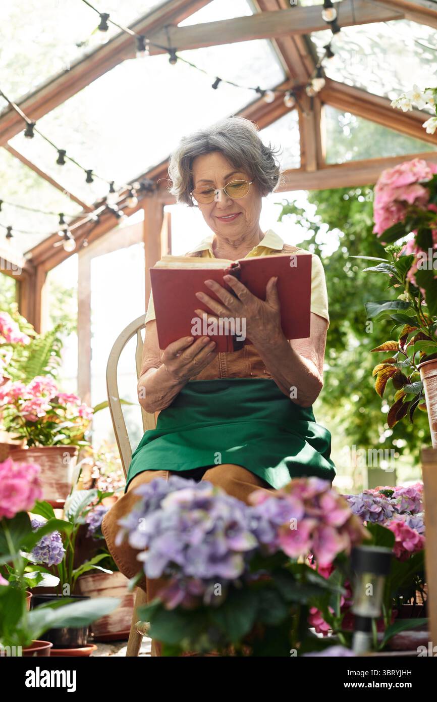 Eine reizende Seniorin liest ein Buch, umgeben von bunten Blumen im Garten. Stockfoto