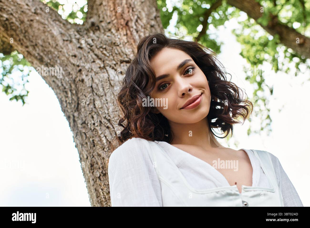 Eine junge Frau mit welligen Haaren lächelt glücklich in einem sonnigen Park, umgeben von üppigem Grün. Stockfoto
