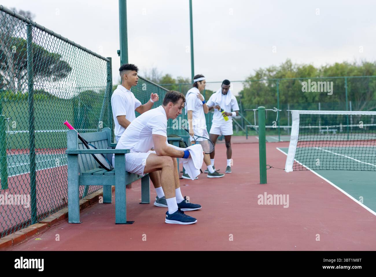 Verschiedene männliche Teamkollegen sitzen und stehen auf dem Tennisplatz und halten Schläger, Handtuch und Wasserflasche Stockfoto