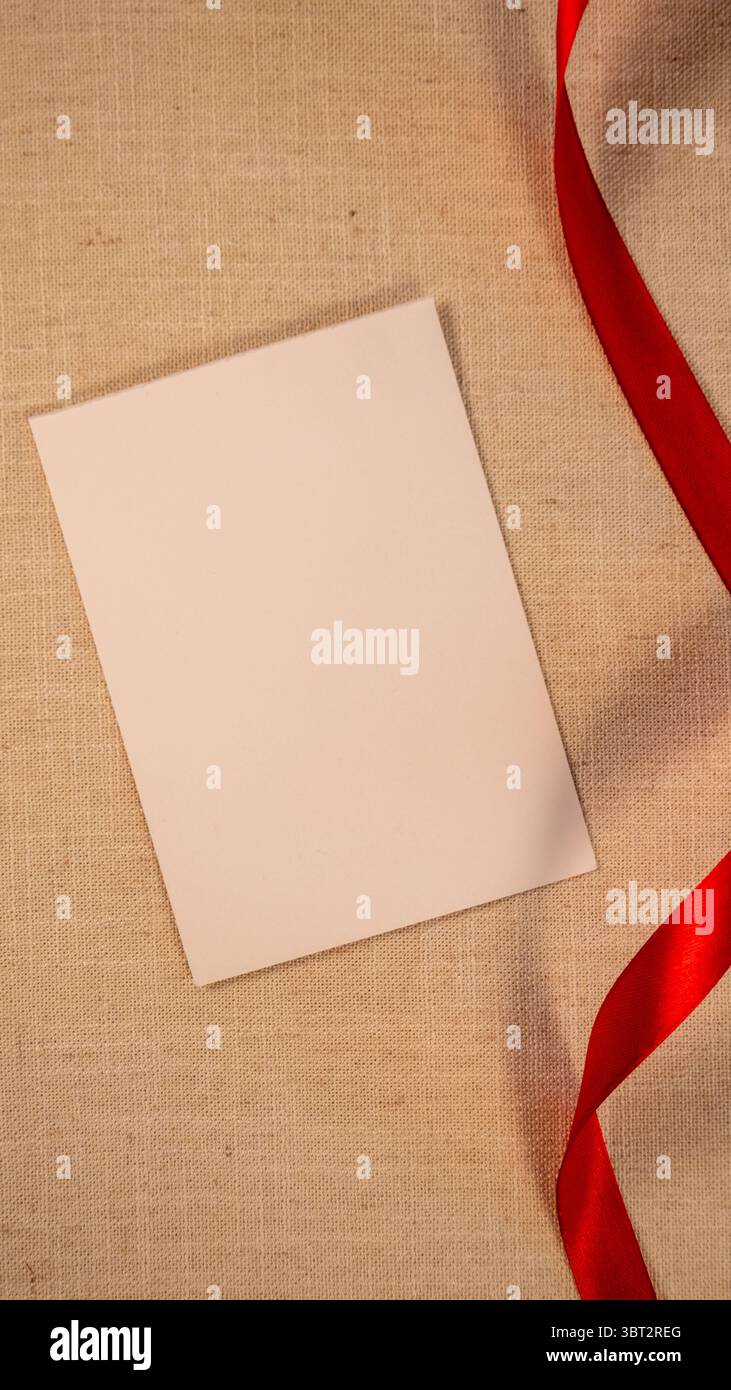 Ansicht von oben: Flache Lage von weißem Grußpapier auf beigefarbenem Stoffhintergrund mit rotem Band, vertikales Foto, Platz für Kopiertext Stockfoto