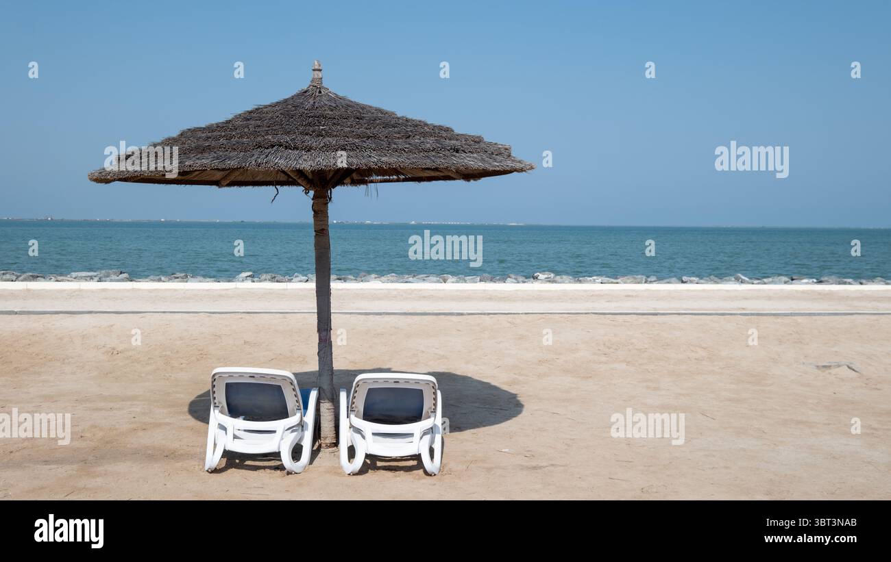 Liegestühle unter einem Strohschirm auf weichem Sand, mit Blick auf das Meer unter einem klaren blauen Himmel, Stockfoto von Entspannen, Sonnenbaden unter Strand um Stockfoto