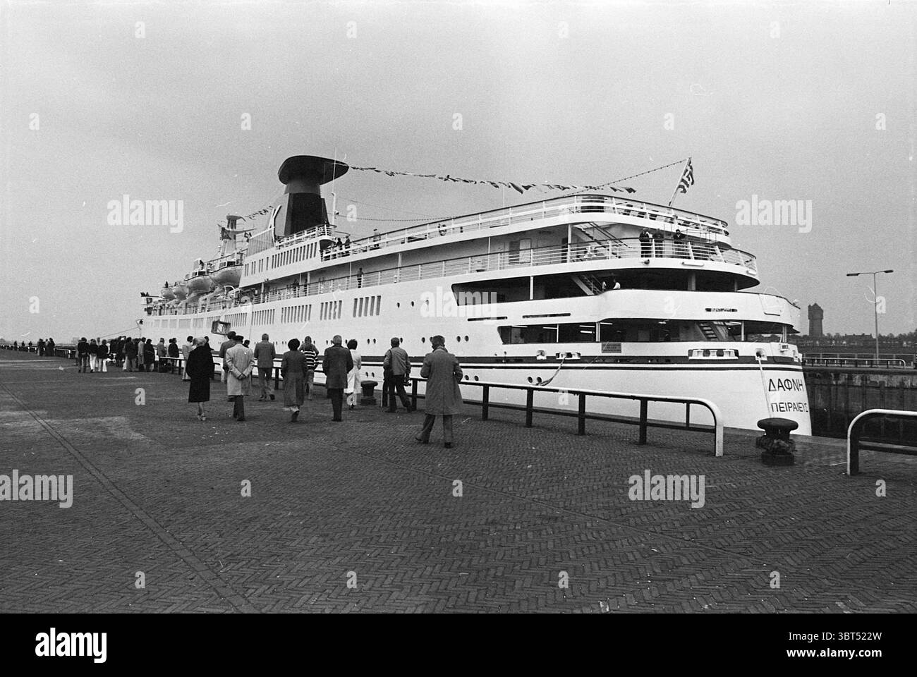 Kreuzfahrtschiff 'The Daphne' Ships, Whizgle News, Dutch Desk, Niederlande, 1950 - 2000 am 28-05-1976. Dies sind die Elemente im Bild. In der Szene legt ein großes, imposantes Schiff an einem Hafen an, dessen schlankes Design einen überwiegend weißen Rumpf mit schwarzen und blauen Elementen zeigt. Das Schiff verfügt über mehrere Decks, deutlich sichtbare Strukturen, die ein gut gestaltetes und geräumiges Inneres suggerieren, das durch Fensterreihen hervorgehoben wird, die den gedämpften Himmel reflektieren. Auf dem Dock sind mehrere Figuren versammelt, die in Mänteln gekleidet sind, die ein Gefühl von Kälte in der Luft vermitteln, was wahrscheinlich auf das Wetter hinweist. Sie Stockfoto