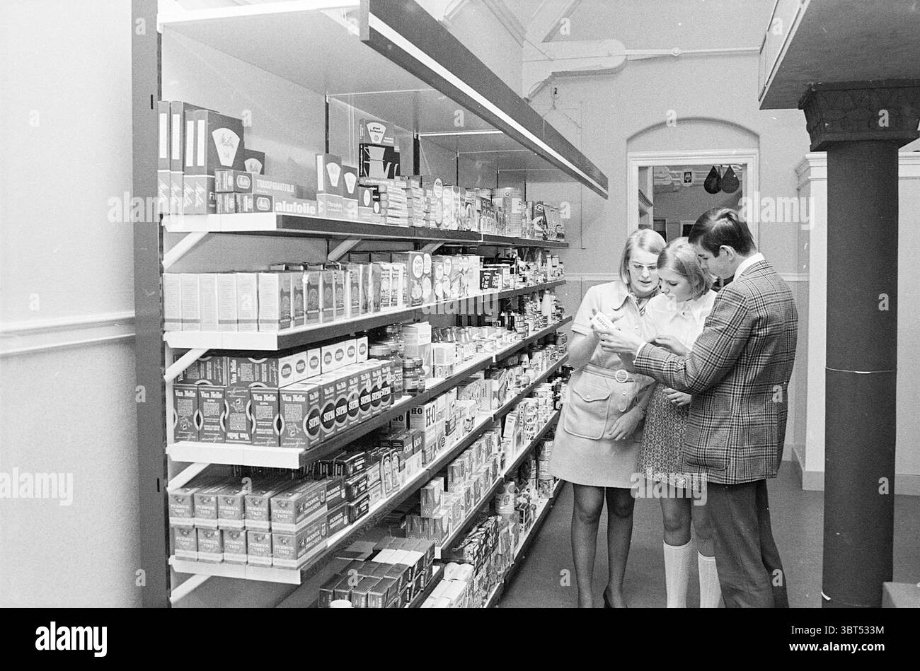 Neues Klassenzimmer als Shop Schulen Haarlem Jacobstraat Niederlande, Whizgle News, Dutch Desk, Niederlande, 1950 - 2000 am 09-12-1970. Diese Themen sind in der Abbildung dargestellt. In einem gut beleuchteten Lebensmittelladen stehen zwei Frauen und ein Mann nahe beieinander, um sich zu unterhalten. Die Frauen tragen leichte Sommeroutfits; eine trägt einen kurzen Rock mit einem taillierten Oberteil, die andere trägt einen ähnlichen Stil, aber in etwas anderer Farbe. Der Mann trägt einen gemusterten Blazer und präsentiert ein poliertes Aussehen. Die Szene ist gesäumt von ordentlich angeordneten Regalen, gefüllt mit einer Reihe von verpackten Waren. T Stockfoto