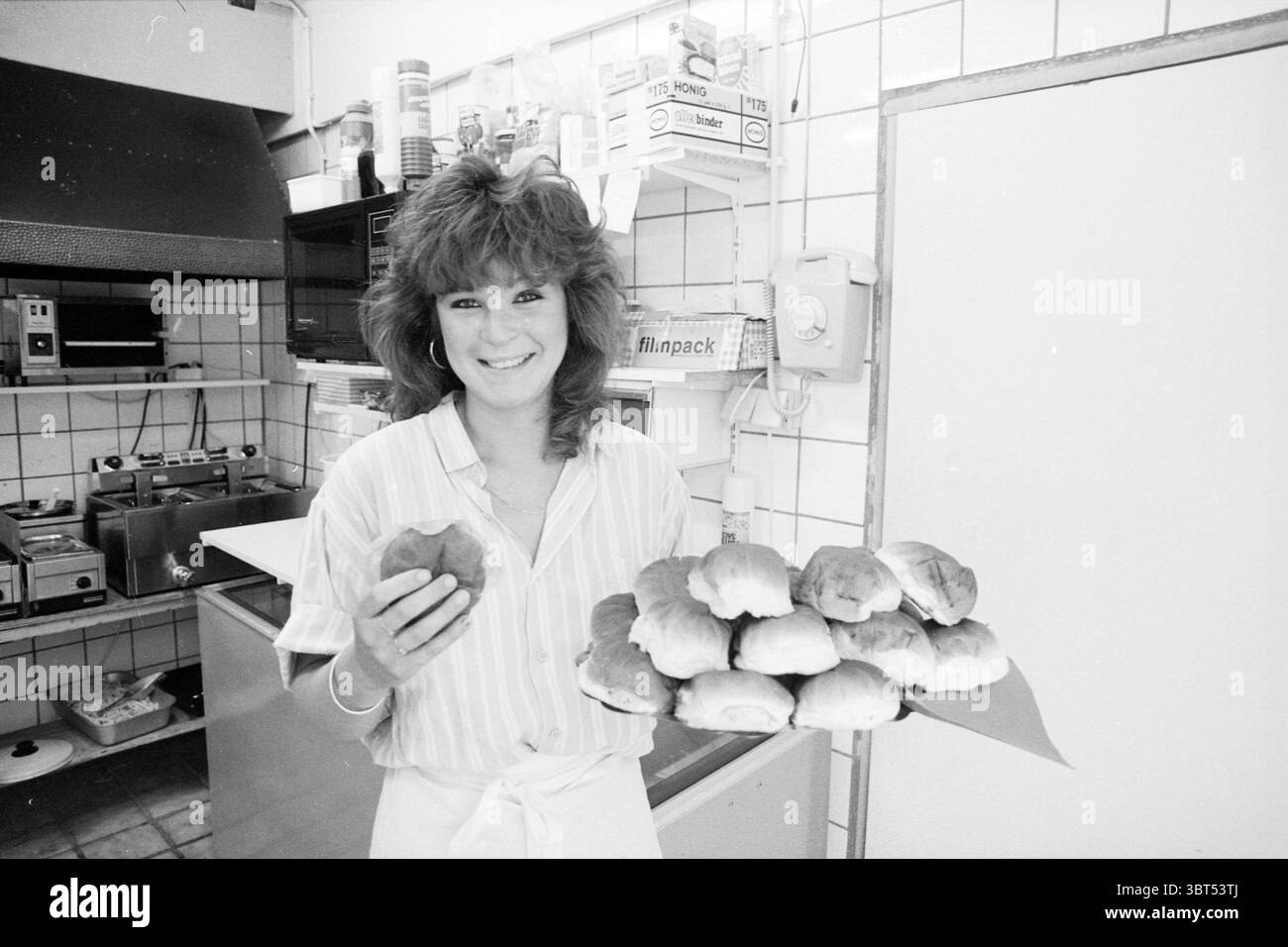 Marchel Flowers Providing Sandwiches Bolletje Eatery IJmuiden Brood IJmuiden the Netherlands, Whizgle News, Dutch Desk, Niederlande, 1950 - 2000 am 15-08-1988. Dies sind die Elemente im Bild. Die Szene zeigt eine fröhliche Person, die in einer hellen und gut beleuchteten Küchenumgebung steht. Sie hat voluminöses, welliges Haar, das ihr Gesicht umrahmt und ein freundliches und einladendes Verhalten ausstrahlt. In der einen Hand hält sie eine einzelne Rolle, während ihr anderer Arm nach außen reicht und eine Platte präsentiert, die mit mehreren ähnlich gebackenen Waren gefüllt ist – goldbraune Brötchen, die ein warmes und appetitliches Aussehen verleihen. Die Stockfoto