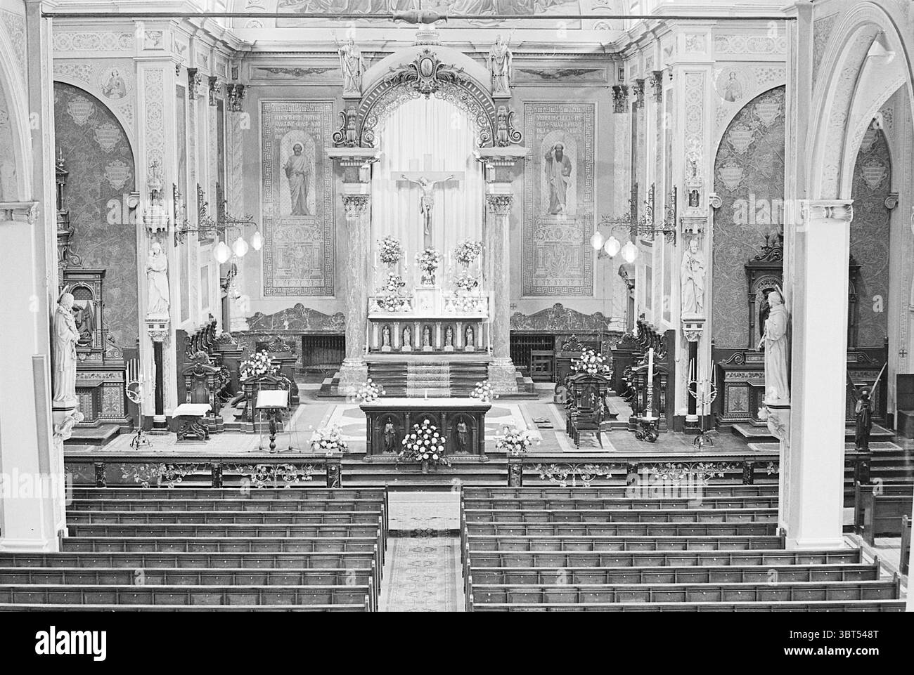 Innenraum einer römisch-katholischen Kirche, Whizgle News, Dutch Desk, Niederlande, 1950 - 2000 im Jahr 1968. Das Bild enthält diese Themen. Die Szene entfaltet sich in einem großen, reich verzierten Inneren einer Kirche, die durch hohe Decken und komplexe architektonische Details gekennzeichnet ist. Der Raum wird überwiegend durch sanfte, atmosphärische Beleuchtung beleuchtet, die von eleganten Armaturen ausgeht und ein warmes Licht auf die polierten Holzoberflächen ausstrahlt. Im Mittelpunkt steht ein wunderschön geschmückter Altar, reich an dekorativen Elementen wie kunstvollen Schnitzereien und Blumenarrangements, die dem Anderen einen Hauch von Farbe verleihen Stockfoto