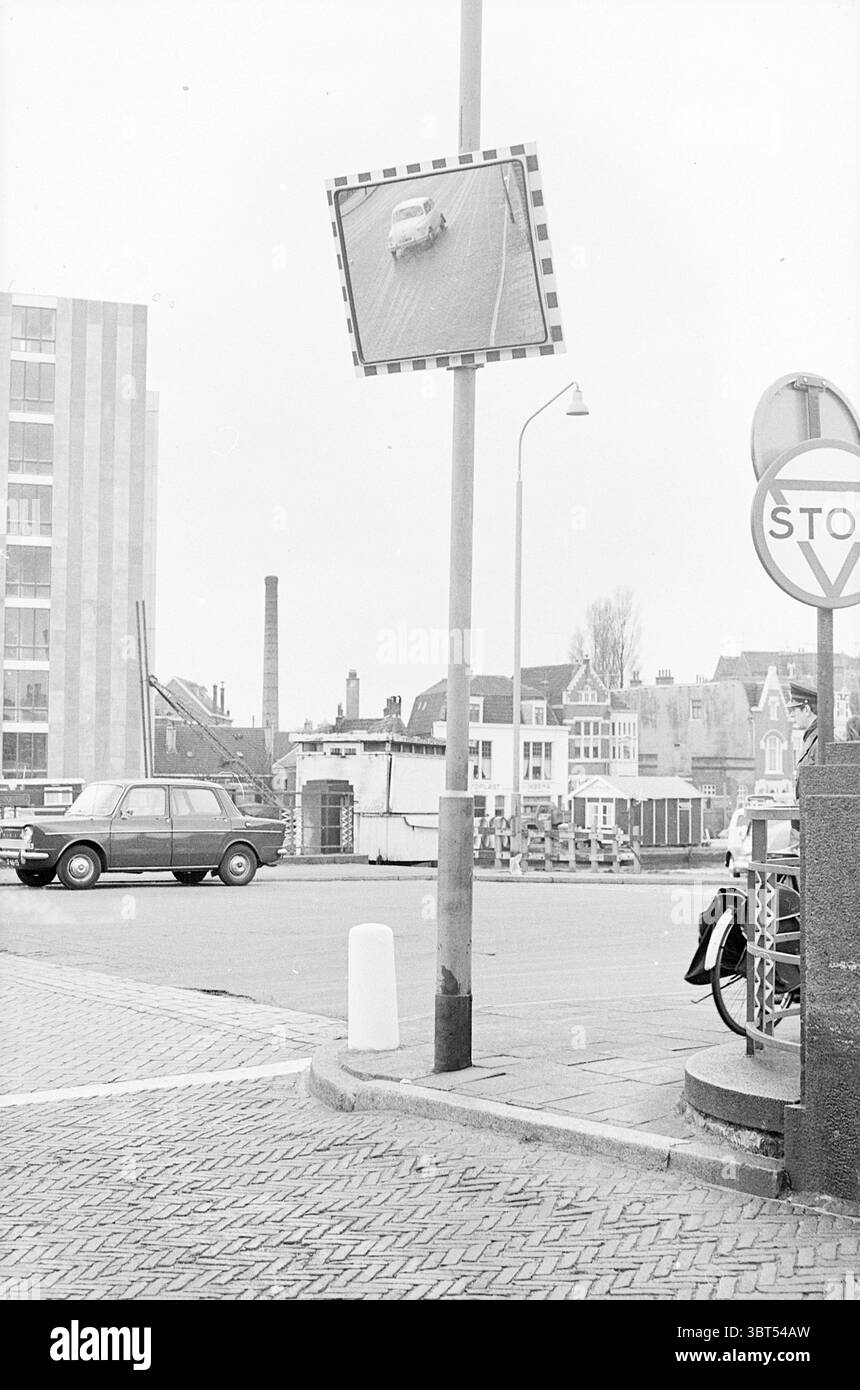 Verkehrsspiegel Herensingel Verkehr sicherer Verkehr Haarlem Herensingel Niederlande, Whizgle News, Dutch Desk, Niederlande, 1950 - 2000 am 29-03-1967. Dies sind die Elemente im Bild. Die Szene fängt eine urbane Umgebung mit einer monochromatischen Palette ein, die stark auf Grautöne ausgerichtet ist. Hervorzuheben ist ein Verkehrsspiegel an einem hohen Pfosten, der die Sicht eines Fahrzeugs auf einer belebten Straße reflektiert. Der Spiegel selbst ist mit kräftigen schwarz-weißen Streifen umrandet, die seine Sichtbarkeit verbessern. Im Hintergrund gibt es verschiedene Gebäude, die eine Mischung aus architektonischem styl vermuten lassen Stockfoto