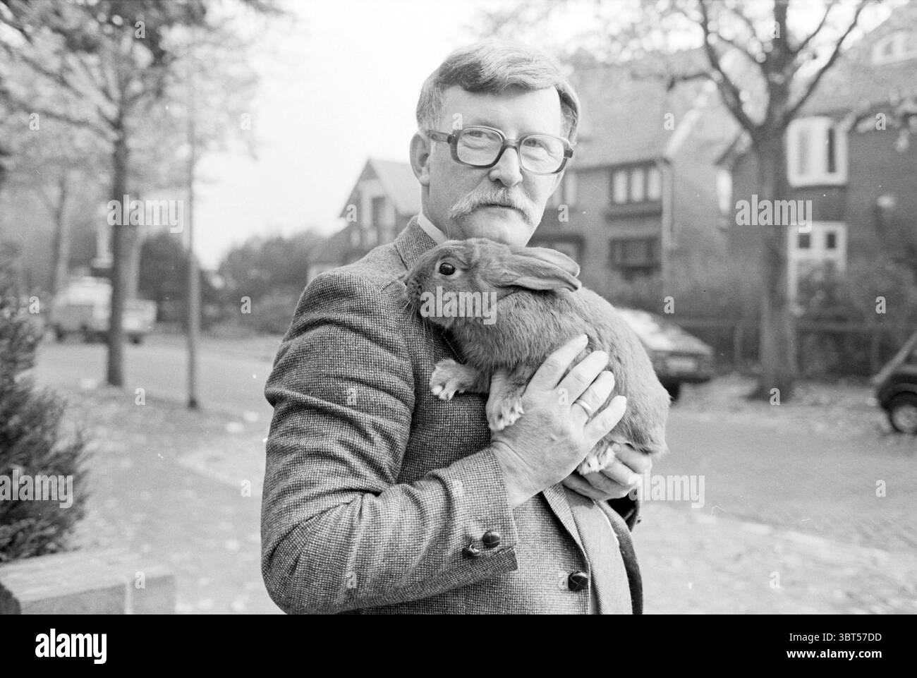 Porträt Mr. Swallow fotografiert auf der Straße mit einem Kaninchen in der Hand, Whizgle News, Dutch Desk, Niederlande, 1950 - 2000 am 28. 10. 1982. Diese Themen sind in der Abbildung dargestellt. In einer Schwarzweißszene steht ein Mann draußen und wiegt ein großes Kaninchen an seiner Brust. Er hat einen sanften Ausdruck, eingerahmt von einer runden Brille und einem markanten Schnurrbart. Seine Kleidung besteht aus einem Tweed-Blazer über einem hellen Hemd, was ihm ein klassisches, zeitloses Aussehen verleiht. Der Hintergrund zeigt eine Vorstadtstraße, die von malerischen Häusern gesäumt ist, mit spitzen Dächern und sichtbaren Details wie Fenstern und Glockenspiel Stockfoto