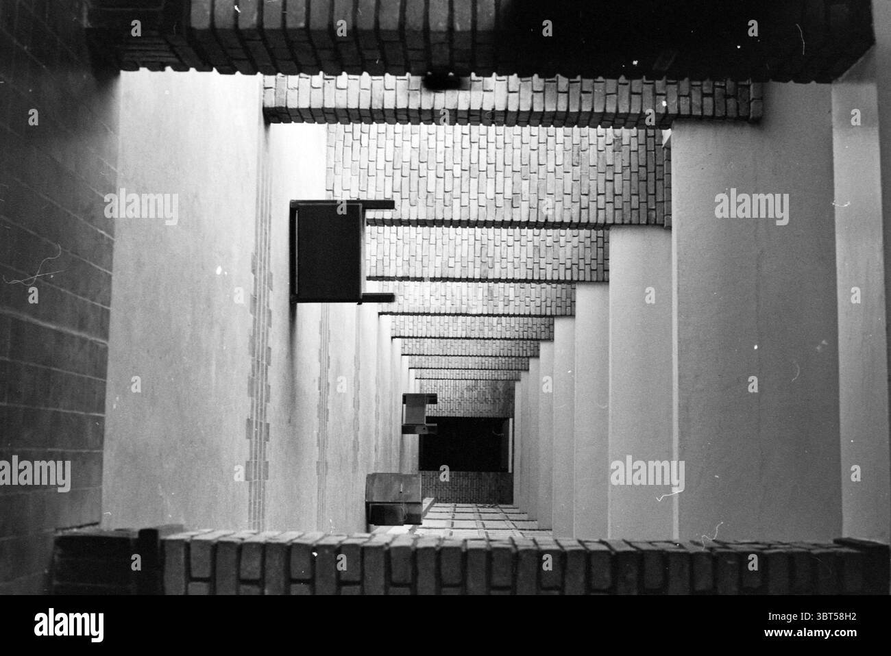 Krematorium Westerveld Driehuis, Whizgle News, Dutch Desk, Niederlande, 1950 - 2000 am 17-04-1991. Diese Themen werden in der Abbildung angezeigt. Die Szene zeigt eine beeindruckende Perspektive eines langen, schmalen Korridors, der durch seine vertikale Ausrichtung gekennzeichnet ist, die den Blick nach unten in einen tiefen Fluchtpunkt zieht. Die Wände sind mit einer Reihe von Backsteinstrukturen verziert, deren Texturen und Muster ein robustes und dennoch klassisches Feeling verleihen. Die Ziegelsteine, die in Grautönen variieren, erzeugen einen optisch anregenden Kontrast zu den weicheren, gedämpften Tönen der glatten weißen Wände. Während sich der Korridor ausdehnt, Schatten Stockfoto