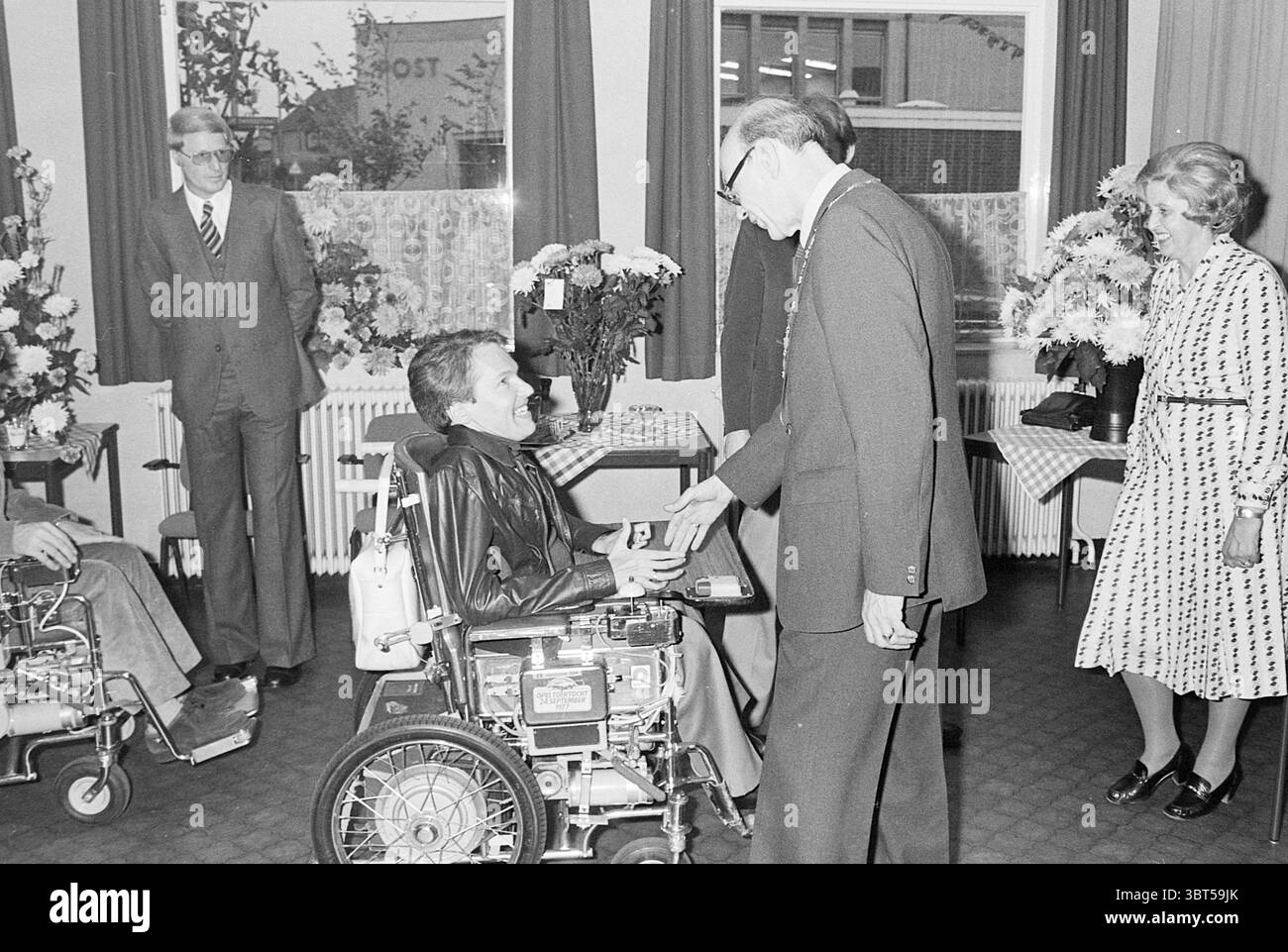 Installation von Bürgermeister Machielsen Zandvoort Mayors, Whizgle News, Dutch Desk, Niederlande, 1950 - 2000 am 05-10-1977. Das Bild enthält diese Themen. In der Szene interagiert eine Gruppe von vier Personen innerhalb eines gut beleuchteten Raumes, dessen Wände mit Dekorationen und Topfblumen geschmückt sind, die trotz der Grautöne ein Gefühl von Wärme vermitteln. Der Boden ist strukturiert, möglicherweise gekachelt oder gemustert, was zu einer strukturierten und dennoch einladenden Umgebung beiträgt. In der Mitte greift ein Mann im Rollstuhl mit einer anderen Person in Kontakt, die in der Nähe steht. Die sitzende Person wirkt entspannt, in dunkler Kleidung gekleidet Stockfoto