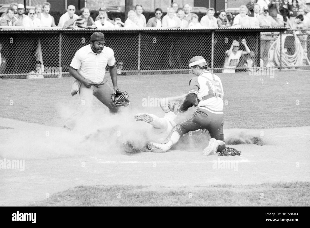 Kinheim - HCAW Baseball Kinheim, Whizgle News, Dutch Desk, Niederlande, 1950 - 2000 auf 30-07-1988. Dies sind die Elemente im Bild. Die Szene fängt einen intensiven Moment auf einem Baseballfeld ein, voller Action und Spannung. Im Vordergrund stehen zwei Spieler im Mittelpunkt der Aufmerksamkeit. Ein Spieler, der in einer leuchtend roten Uniform mit markanten weißen Streifen gekleidet ist, rutscht in die Startplatte und schleudert eine Staubwolke um ihn herum. Sein Körper ist tief am Boden positioniert und zeigt Leichtathletik und Dringlichkeit. Der Fänger, gekleidet in einer kontrastierenden hellblauen Uniform, ist tief gehockt, voll eingekuppelt Stockfoto