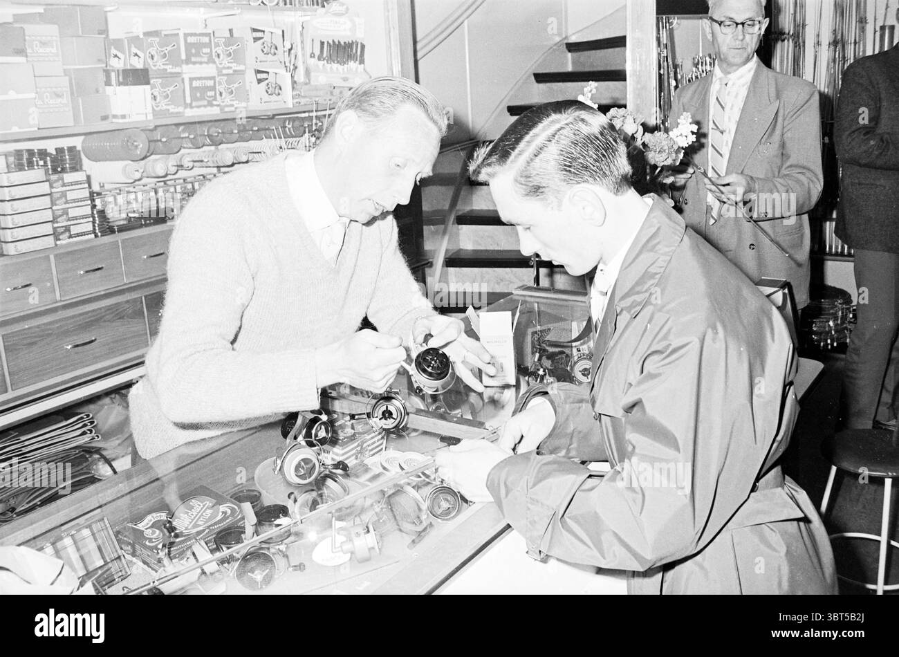 Fisherman's Shop, Whizgle News, Dutch Desk, Niederlande, 1950 - 2000 am 05-1961. Diese Themen sind in der Abbildung dargestellt. In einem gut beleuchteten Geschäft sind zwei Personen an einer Transaktion beteiligt, an einer Glasvitrine, die mit verschiedenen Gegenständen überfüllt ist. Ein Mann, in einem hellen Pullover gekleidet, beugt sich mit einem konzentrierten Blick über die Theke und untersucht ein Objekt genau. Sein Verhalten deutet darauf hin, dass er sachkundig und aufmerksam ist und vielleicht versucht, einen Verkauf zu tätigen oder Ratschläge anzubieten. Der zweite Mann, in einem dunklen, glänzenden Mantel gekleidet, steht ihm gegenüber und hält einen Gegenstand in der Hand. Sein Ausdruck ist neugierig Stockfoto