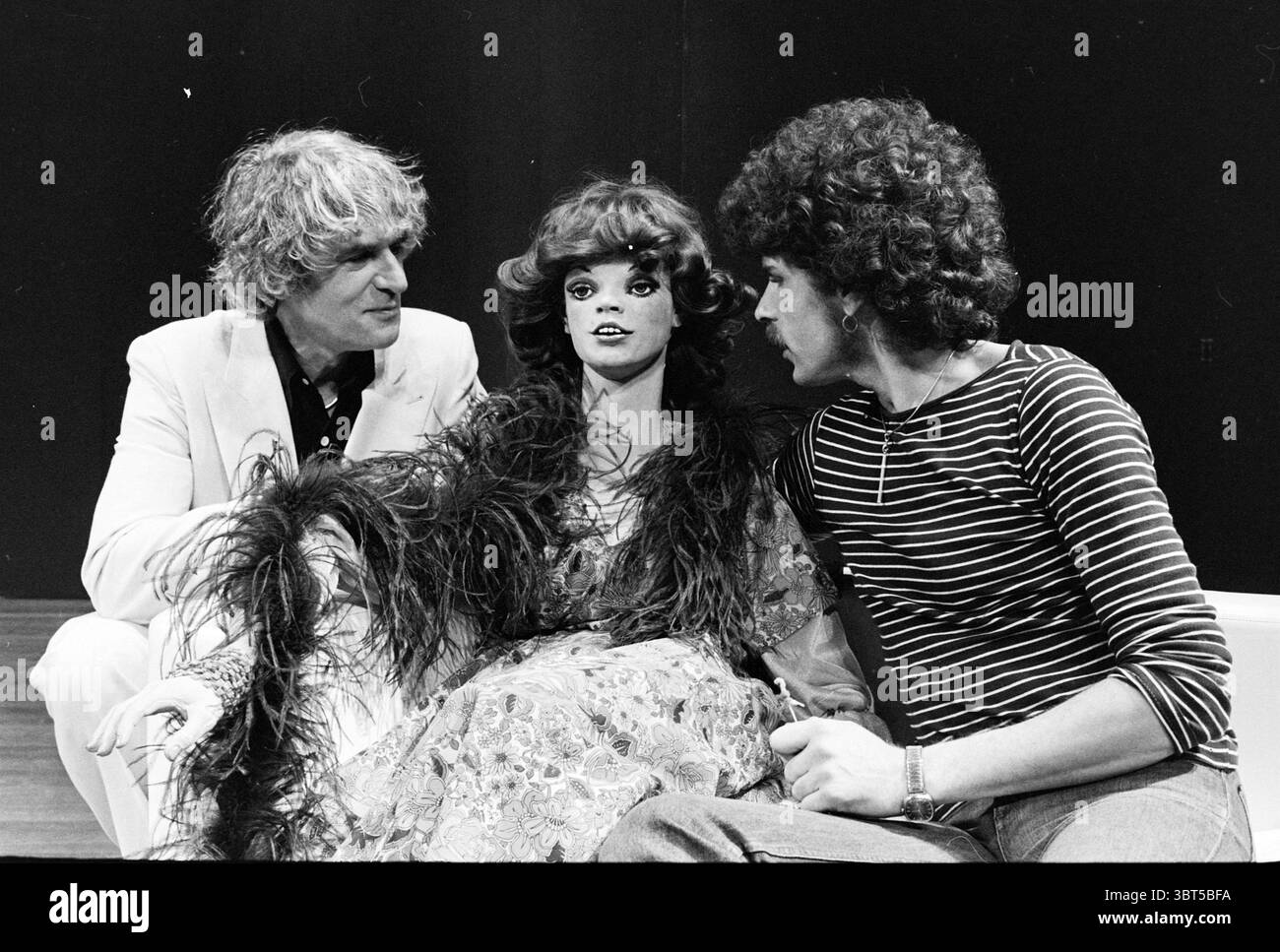 R. Long und D. Frenkel Frank „What Every Girl should Know“ Robert Long, Whizgle News, Dutch Desk, Niederlande, 1950 - 2000 am 07-11-1977. Dies sind die Themen im Bild. Die Szene zeigt einen auffälligen Kontrast zwischen drei eng aneinander sitzenden Figuren. Auf der einen Seite trägt ein Mann mit hellen, lockigen Haaren einen eleganten weißen Anzug, der einen Hauch von Raffinesse ausstrahlt. Seine Haltung ist entspannt, aber aufmerksam, was auf ein Gefühl der Bindung an die anderen hindeutet. Zwischen ihm und einer anderen Person sitzt eine Schaufensterpuppe oder eine Puppe, die mit dramatischem Make-up, langen welligen Haaren und einem bunten, Stockfoto