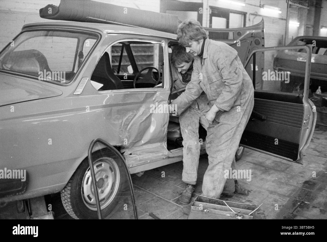 Zwei Mechaniker in einer Garage mit beschädigtem Auto, Whizgle News, Dutch Desk, Niederlande, 1950 - 2000 am 29.01.1979. Dies sind die Themen im Bild. Die Szene zeigt zwei Personen, die in einer schwach beleuchteten Garage an einem Oldtimer arbeiten. Die vorherrschende Farbe des Fahrzeugs ist ein gedämpfter, leicht verblichener Rotton, der sein Alter unterstreicht. Das Fahrzeug hat sichtbare Schäden entlang der Beifahrerseite, wo das Metall zerknittert und freigelegt ist. Eine Person lehnt sich von der Beifahrerseite in das Auto, während die andere über die offene Tür neigt, beide mit zweckmäßigen Overalls verkleidet, die mit Fett und d gefärbt sind Stockfoto