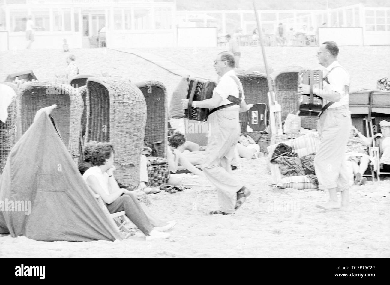 Menschen an einem Tag am Strand Menschen und Dinge Strand und Strände, Whizgle News, Dutch Desk, Niederlande, 1950 - 2000 am 04-08-1963. Dies sind die Elemente im Bild. Die Szene fängt eine lebhafte Strandkulisse mit Menschen ein, die einen sonnigen Tag am Ufer genießen. Mehrere Personen sind über die Sandlandschaft verstreut und zeigen eine Reihe von Aktivitäten. Links sitzt eine Frau bequem in einem zusammenklappbaren Stuhl, in ihrer Umgebung, während rechts zwei Männer sich beim Akkordeon abheben und der Szene eine festliche Atmosphäre verleihen. Die beiden Musiker sind lässig gekleidet, Ref Stockfoto