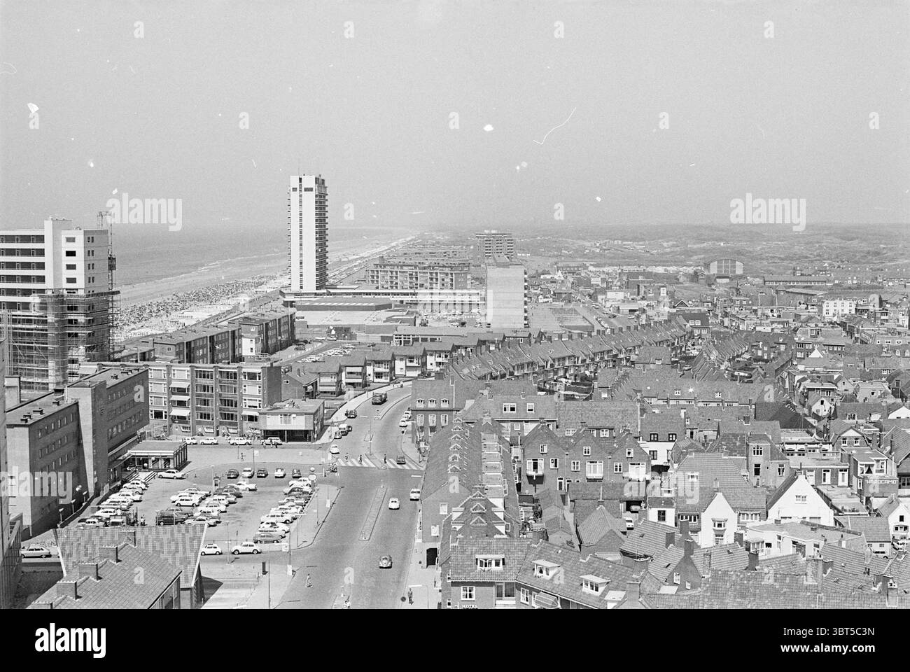 Übersicht Zandvoort Zandvoort, Whizgle News, Dutch Desk, Niederlande, 1950 - 2000 am 04-07-1969. Das Bild enthält diese Themen. Die Szene zeigt eine weite Luftaufnahme einer Küstenstadt mit einer Mischung aus urbanen und natürlichen Elementen. Im Vordergrund zeigt eine Reihe dicht gepackter Wohngebäude einen charakteristischen architektonischen Stil mit Giebeldächern, höchstwahrscheinlich aus Ziegeln oder Fliesen, die ein strukturiertes Muster gegen den Himmel schaffen. Die Gebäude variieren in Höhe und Design, was der Komposition Tiefe verleiht. In Richtung Mittelteil schlängelt sich die Straße durch die Stadtlandschaft, Stockfoto