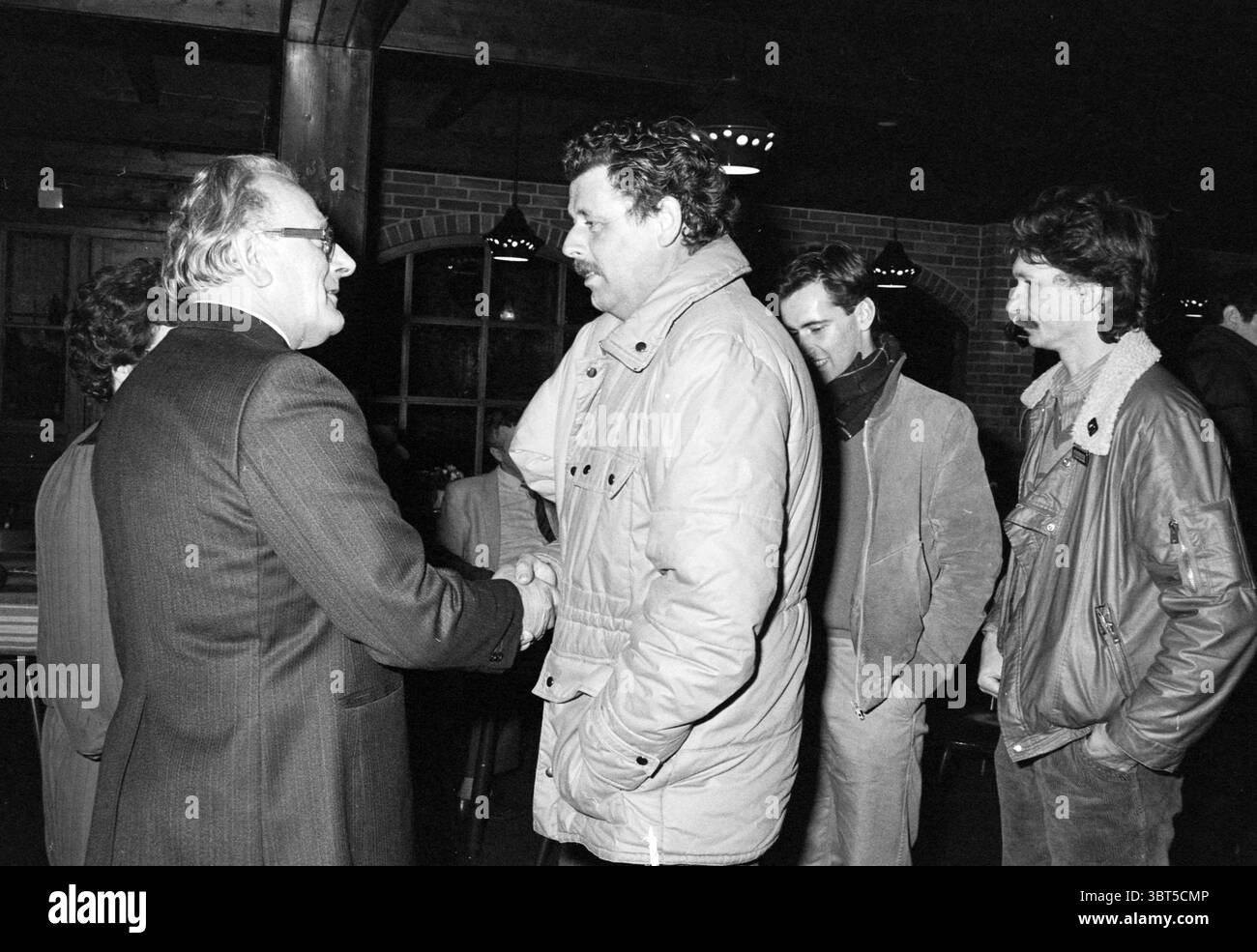Abschiedsempfang Mr. Blokker HD Haarlems Dagblad etc. Und IJmuider Courant and Koeri, Whizgle News, Dutch Desk, Niederlande, 1950 - 2000 am 14-02-1986. Dies sind die Themen im Bild. In einem schwach beleuchteten Innenraum führt eine Gruppe von Einzelpersonen Gespräche und schafft eine Szene, die Wärme mit einem Hauch von Formalität in Einklang bringt. Die Einrichtung verfügt über Holzbalken und eine warme Backsteinkulisse, die zu einem einladenden Ambiente beiträgt. An vorderster Front schütteln zwei Männer die Hand. Einer, der in einem maßgeschneiderten dunklen Anzug gekleidet ist, strahlt einen Hauch von Professionalität und Selbstvertrauen aus. Der andere trägt Stockfoto