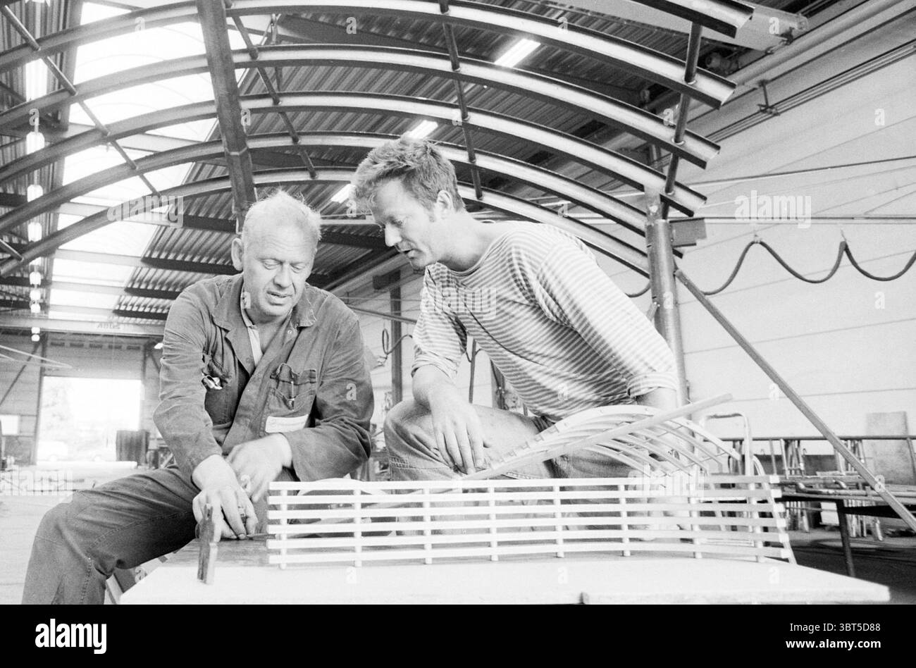 Kunstwerk bei der Firma de Groot Heemskerk Heemskerk, Whizgle News, Dutch Desk, Niederlande, 1950 - 2000 am 02.09.1991. Das Bild enthält diese Themen. In einer geräumigen Werkstatt führen zwei Männer enge Gespräche über eine auf einer Werkbank aufgestellte Modellstruktur. Die Umgebung zeichnet sich durch industrielle Elemente aus – Stahlträger, die über dem Boden hängen, Schatten und Struktur im offenen Raum bilden. Ein Mann, der in einem Arbeitshemd gekleidet ist, scheint konzentriert zu sein und sich leicht nach vorne zu lehnen, während er das Modell untersucht. Seine Hände, verwittert und stark, zeugen von jahrelanger Handwerkskunst. Ihm gegenüber, der zweite Mann, Stockfoto
