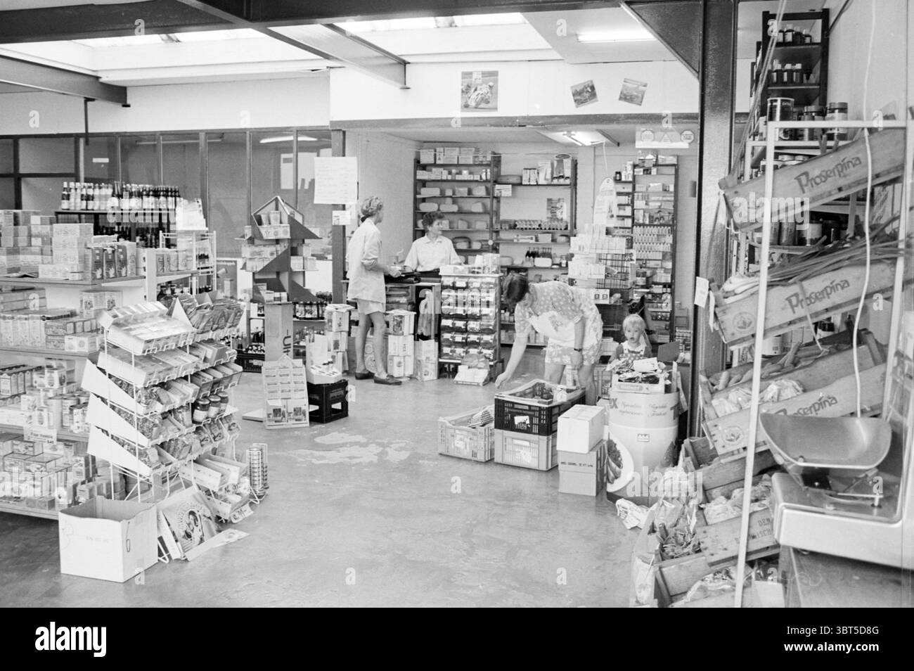 Lebensmittelgeschäft IJm. IJmuiden the Netherlands, Whizgle News, Dutch Desk, Niederlande, 1950 - 2000 am 01-06-1993. Diese Themen sind in der Abbildung dargestellt. Die Szene zeigt einen überdachten Markt oder ein kleines Lebensmittelgeschäft mit einer lebhaften Atmosphäre. Der Raum wird durch die Deckenbeleuchtung beleuchtet, die einen hellen, aber leicht sanften Glanz im Raum ausstrahlt. Die Wände sind in neutralen Tönen lackiert, wodurch die ausgestellten Produkte hervorstechen. Auf der einen Seite gibt es Regale mit einer Vielzahl von Konserven, Snacks und anderen verpackten Artikeln, die alle in einem etwas organisierten Chaos angeordnet sind, das einen Sinn vermittelt Stockfoto