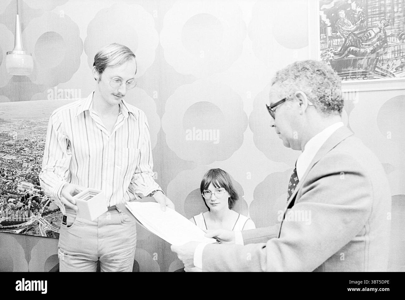 Übergabe von Unterschriften gegen Bauwohnungen Beverwijk Übergabe und Transfer, Whizgle News, Dutch Desk, Niederlande, 1950 - 2000 am 13.06.1977. Dies sind die Elemente im Bild. Die Szene spielt in einem Raum im Retro-Stil, der von großen, kreisförmigen Mustern an den Wänden geprägt ist, die eine lebhafte, aber nostalgische Atmosphäre ausstrahlen. Sanfte Beleuchtung füllt den Raum und schafft eine warme, einladende Stimmung. Im Vordergrund stehen zwei Männer im Zentrum der Komposition. Ein Mann, mit einem schlanken Bauwerk und einer dünnen Brille, steht und hält eine kleine Box, die scheinbar mit Karten oder Folie gefüllt ist Stockfoto