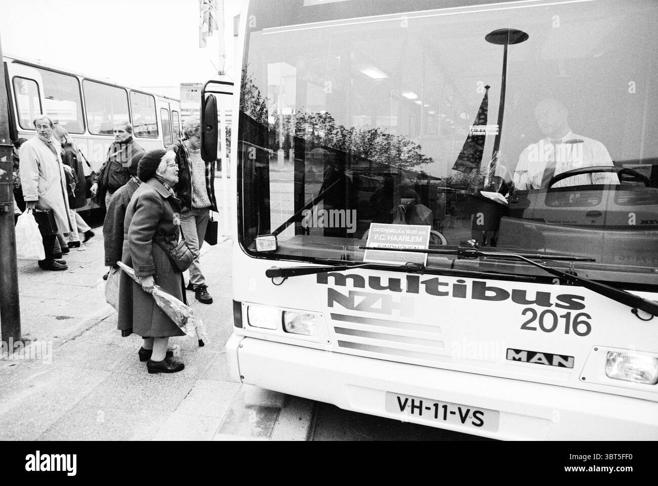 Multibus Schalkwijk Haarlem the Netherlands, Whizgle News, Dutch Desk, Niederlande, 1950 - 2000 auf 31-10-1993. Diese Themen sind in der Abbildung dargestellt. Die Szene fängt einen Moment an einem belebten Busbahnhof ein, reich an menschlicher Interaktion und alltäglichem Leben. Im Vordergrund steht eine ältere Frau mit einem ruhigen, aber neugierigen Ausdruck, ihr Gesicht wird teilweise von einem dunklen Hut und Schal verdeckt. Sie trägt einen langen Mantel, der zu ihrem würdevollen Verhalten beiträgt, während sie auf den Busfahrer blickt, der hinter dem Lenkrad sitzt. Der Bus selbst zeichnet sich durch sein schlankes Design und seine PA aus Stockfoto
