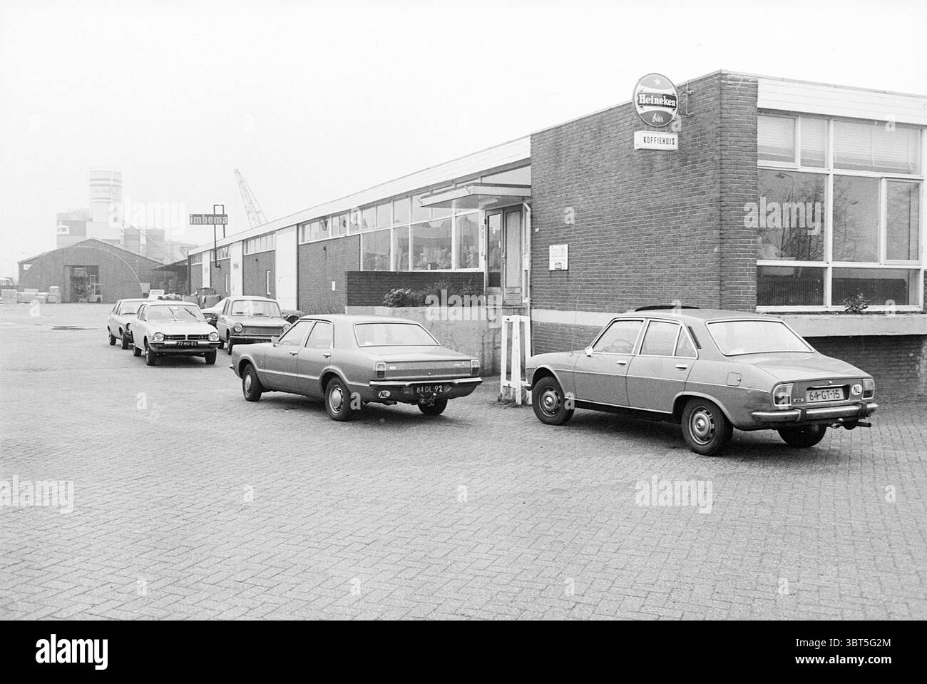 Coffee House, Whizgle News, Dutch Desk, Niederlande, 1950 - 2000 im Jahr 1978. Das Bild enthält diese Themen. Die Szene zeigt ein gedämpftes Industriegebiet, das durch eine Reihe von Fahrzeugen gekennzeichnet ist, die eine gepflasterte Oberfläche säumen. Die Autos, meist Oldtimer, zeigen Grau- und Blautöne, was die düstere Atmosphäre insgesamt noch verstärkt. Ein leicht beschädigtes Gebäude mit großen Fenstern steht auf einer Seite und weist auf seine Funktion als Gewerberaum oder Lagerraum hin. Die Ziegelfassade wird durch eine einfachere Struktur ergänzt, die sich nach außen erstreckt und ein zweckmäßiges Design widerspiegelt. Die Einstellung wird als Overcas angezeigt Stockfoto