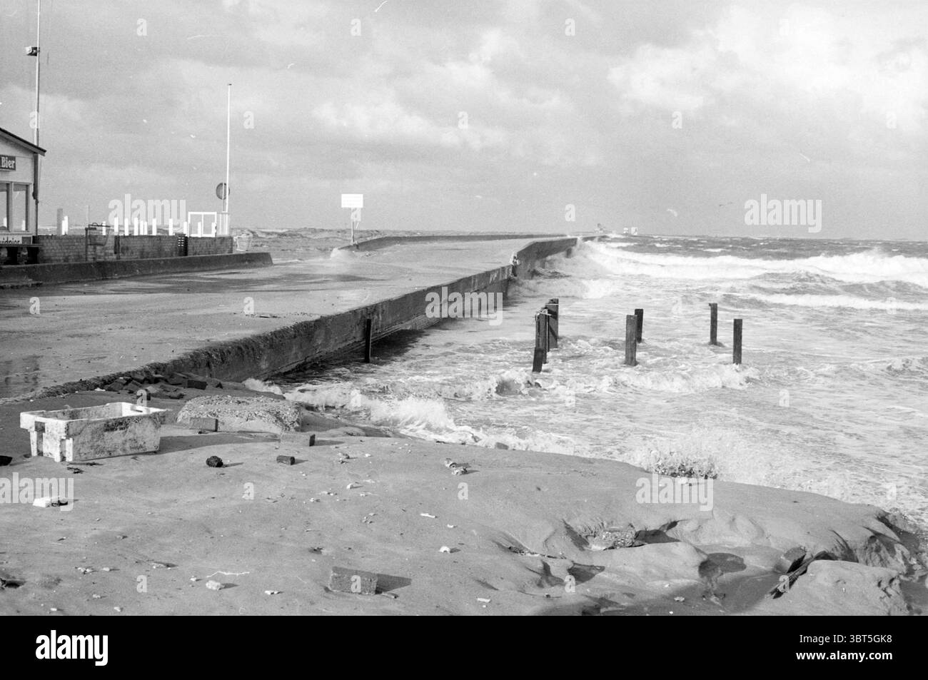 Hochwasserstrand IJmuiden Strand und Strände IJmuiden Niederlande, Whizgle News, Dutch Desk, Niederlande, 1950 - 2000 am 06-11-1985. Das Bild enthält diese Themen. Die Szene zeigt eine Küstenlandschaft unter einem dramatischen Himmel, wo dunkle Wolken über sich heraustreten und auf einen drohenden Sturm hinweisen. Die Küste verfügt über einen robusten, strukturierten Bereich aus nassem Sand, der dank der monochromen Palette eine Vielzahl von Grau- und Weißtönen reflektiert. In die Ferne zieht sich eine breite Betonpromenade, die von einem turbulenten Meer umgeben ist, mit Wellen, die heftig gegen den Rand prallen und Wasser auf die Oberfläche spritzen. Die Stockfoto