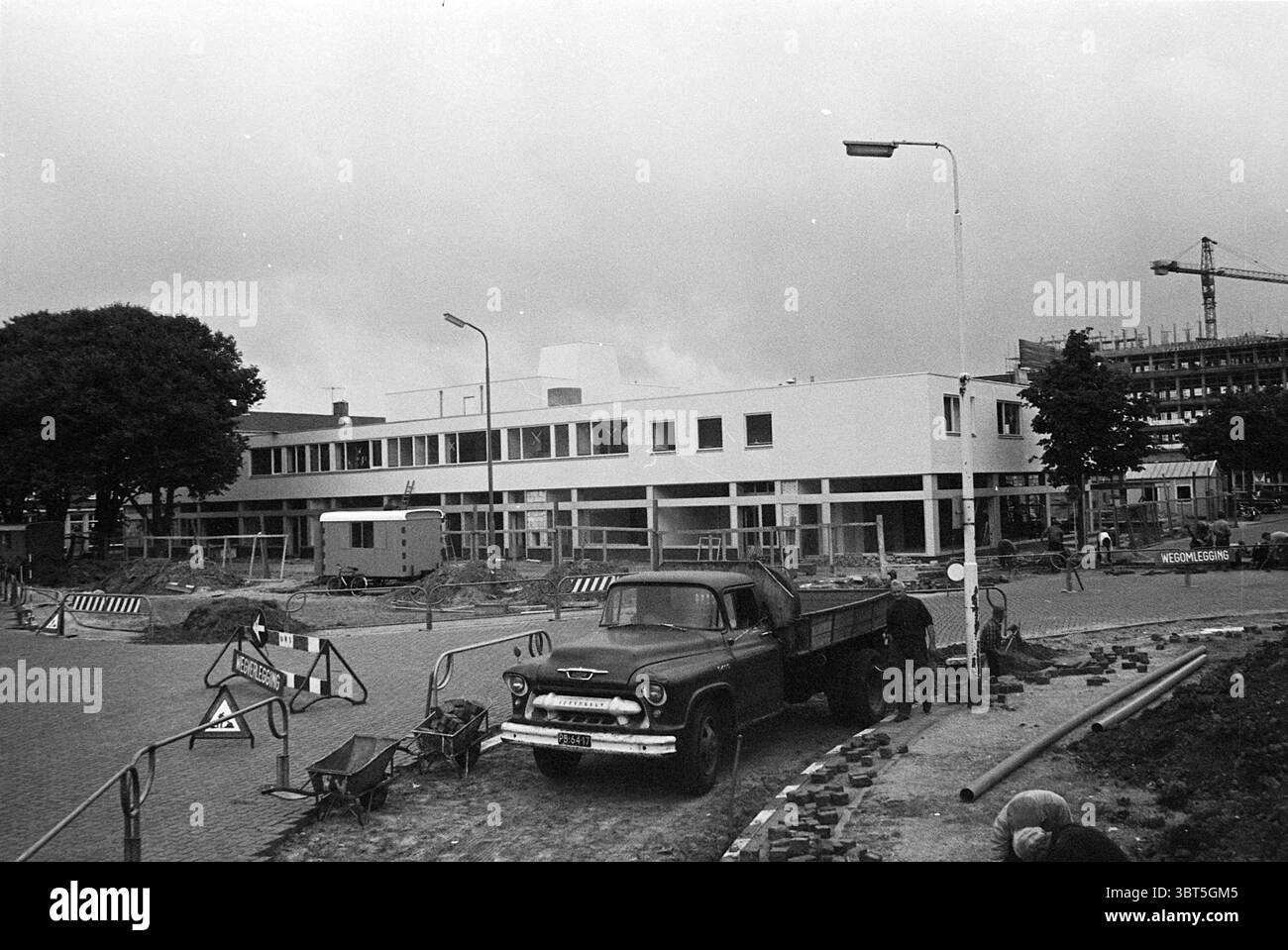 Arbeiten am Marktplein Einkaufszentrum Works, Whizgle News, Dutch Desk, Niederlande, 1950 - 2000 am 25-07-1967. Das Bild enthält diese Themen. Die Szene fängt eine städtische Baustelle mit einer überwiegend monochromatischen Farbpalette ein, die ein Gefühl von Nostalgie weckt. Im Vordergrund steht ein Oldtimer-Lkw, der ein robustes Äußeres und ein industrielles Flair zeigt. Die abgerundeten Linien und die robuste Bauweise des Lkws deuten auf sein Alter und seinen Zweck hin und stehen im Kontrast zu den schlanken Linien des modernen Gebäudes, das im Hintergrund gebaut wird. Links verschiedene Bauschranken, markiert mit ca. Stockfoto