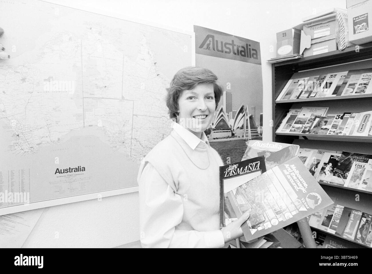 Mrs. V.d. Yard Australian Tourist Board Travel Agencyx, Whizgle News, Dutch Desk, Niederlande, 1950 - 2000 am 28-03-1985. Dies sind die Elemente im Bild. Die Szene zeigt eine Frau, die in einem gut organisierten Raum steht, der mit Reisematerial geschmückt ist. Sie hat einen herzlichen Ausdruck, der auf Zugänglichkeit und Begeisterung hindeutet. Die Frau trägt einen hübschen, hellen Pullover, der einen sanften Kontrast zur dunkleren Umgebung bildet. Im Hintergrund bedeckt eine große Karte von Australien einen Teil der Mauer und bietet einen geografischen Kontext mit gedämpften Farben, die Land und Wasser anzeigen. Ne Stockfoto